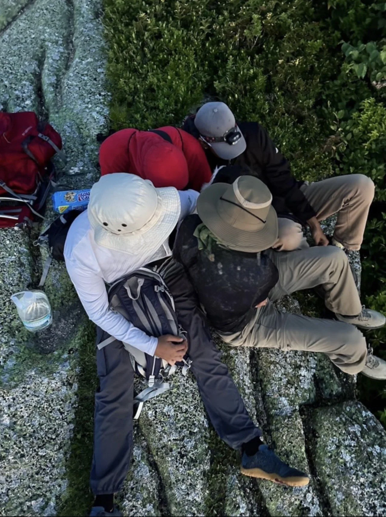 Four people resting on a rocky surface surrounded by green bushes, with backpacks and a water jug nearby, wearing hiking gear including hats, caps, and outdoor clothing.