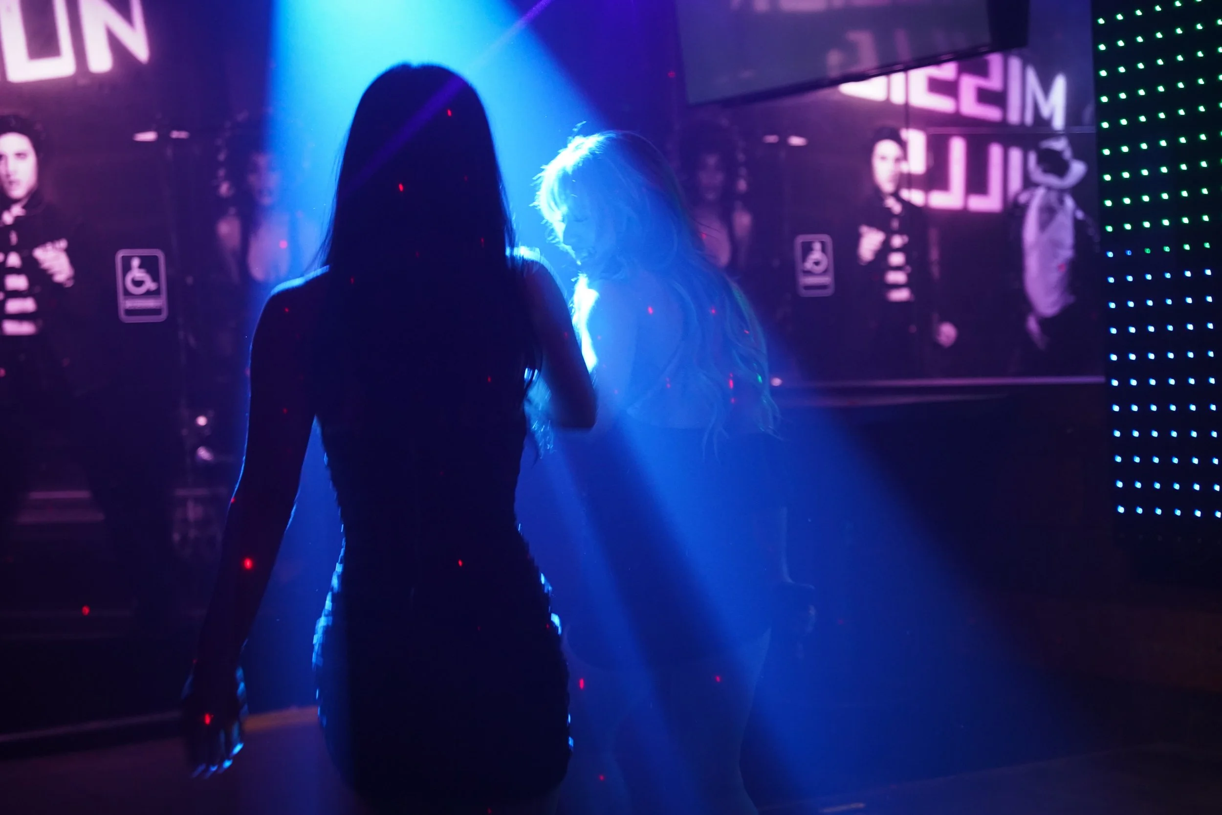 Two women dancing in a nightclub with a backlit DJ booth and colorful lights.