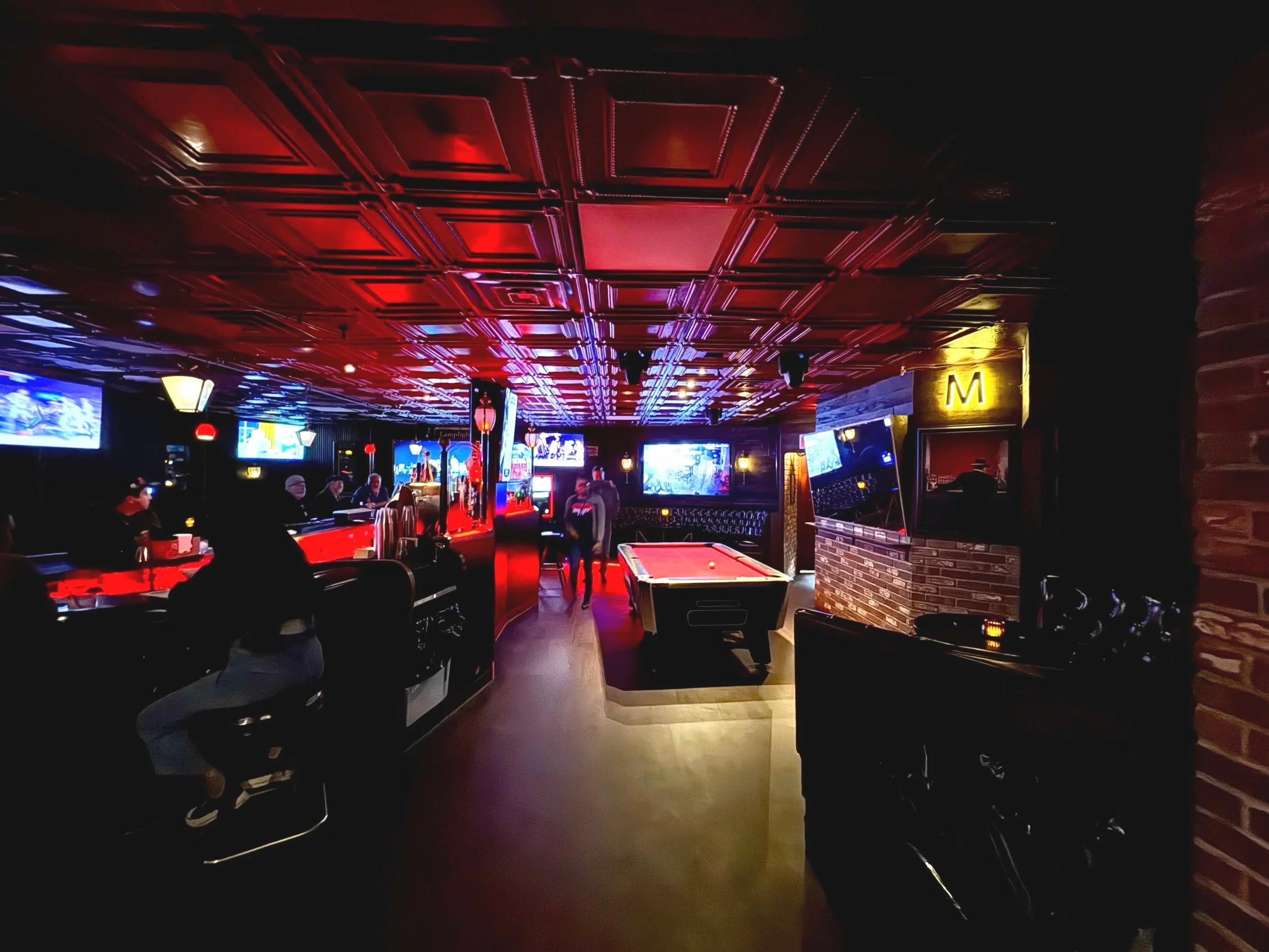 Interior of a dimly lit bar with a pool table in the center, multiple TVs on the walls, and patrons sitting at the bar and tables. Neon lights and a red ceiling create a vibrant atmosphere.