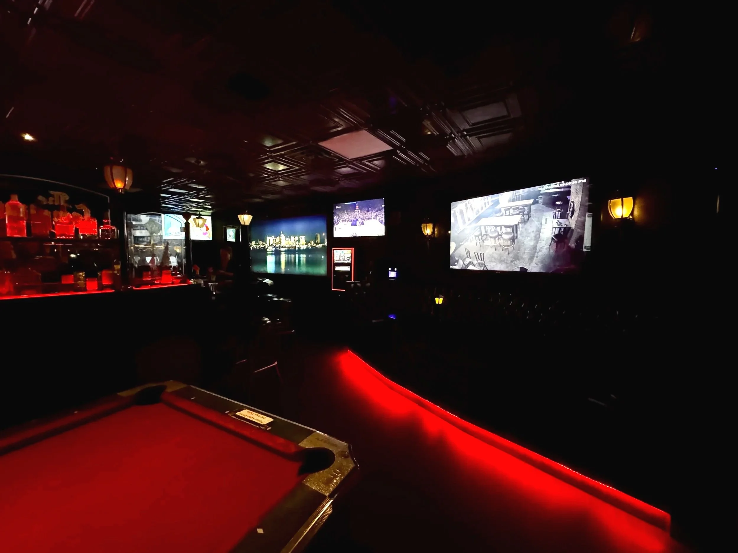 Dark interior of a bar or entertainment venue with multiple large screens displaying different images and a pool table in the foreground, illuminated by warm yellow light fixtures and red accent lighting.