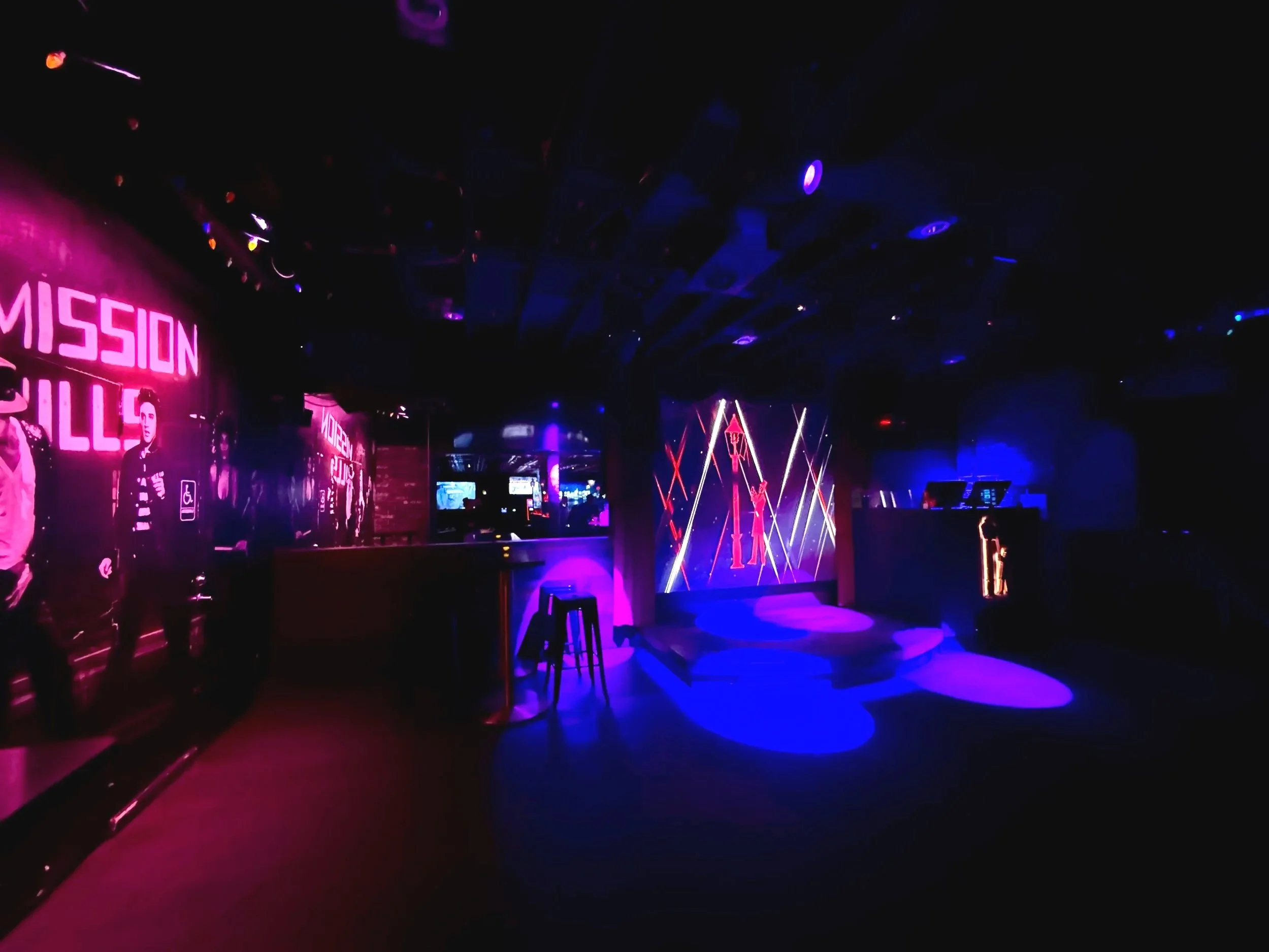 A dimly lit nightclub with a colorful neon stage and LED screens, featuring abstract lighting patterns and people at the bar.