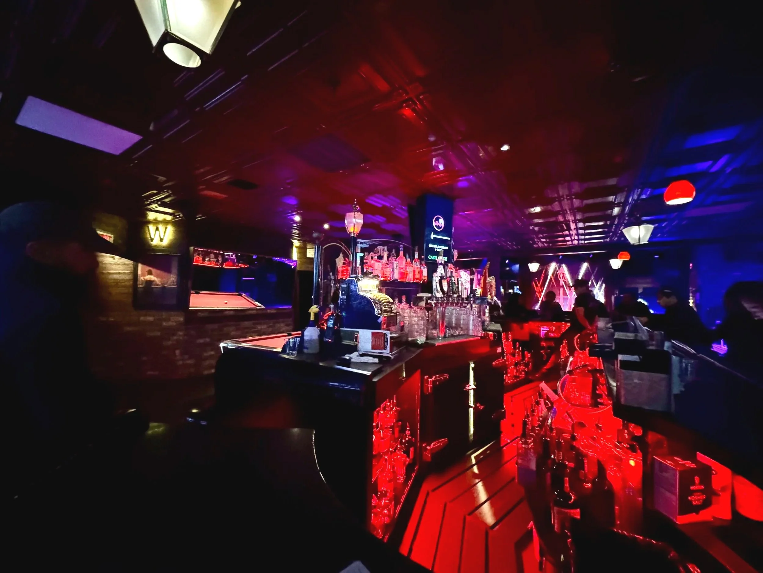 A dimly lit bar with a neon-lit counter and purple and red lighting, with people standing and socializing in the background.