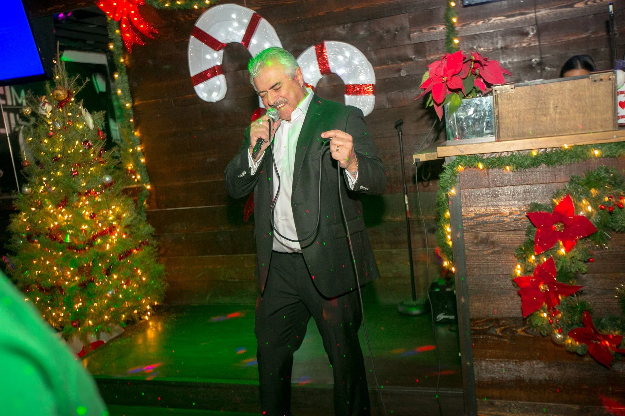 A man in a black suit and white shirt singing into a microphone on a decorated holiday stage with Christmas tree and poinsettias.