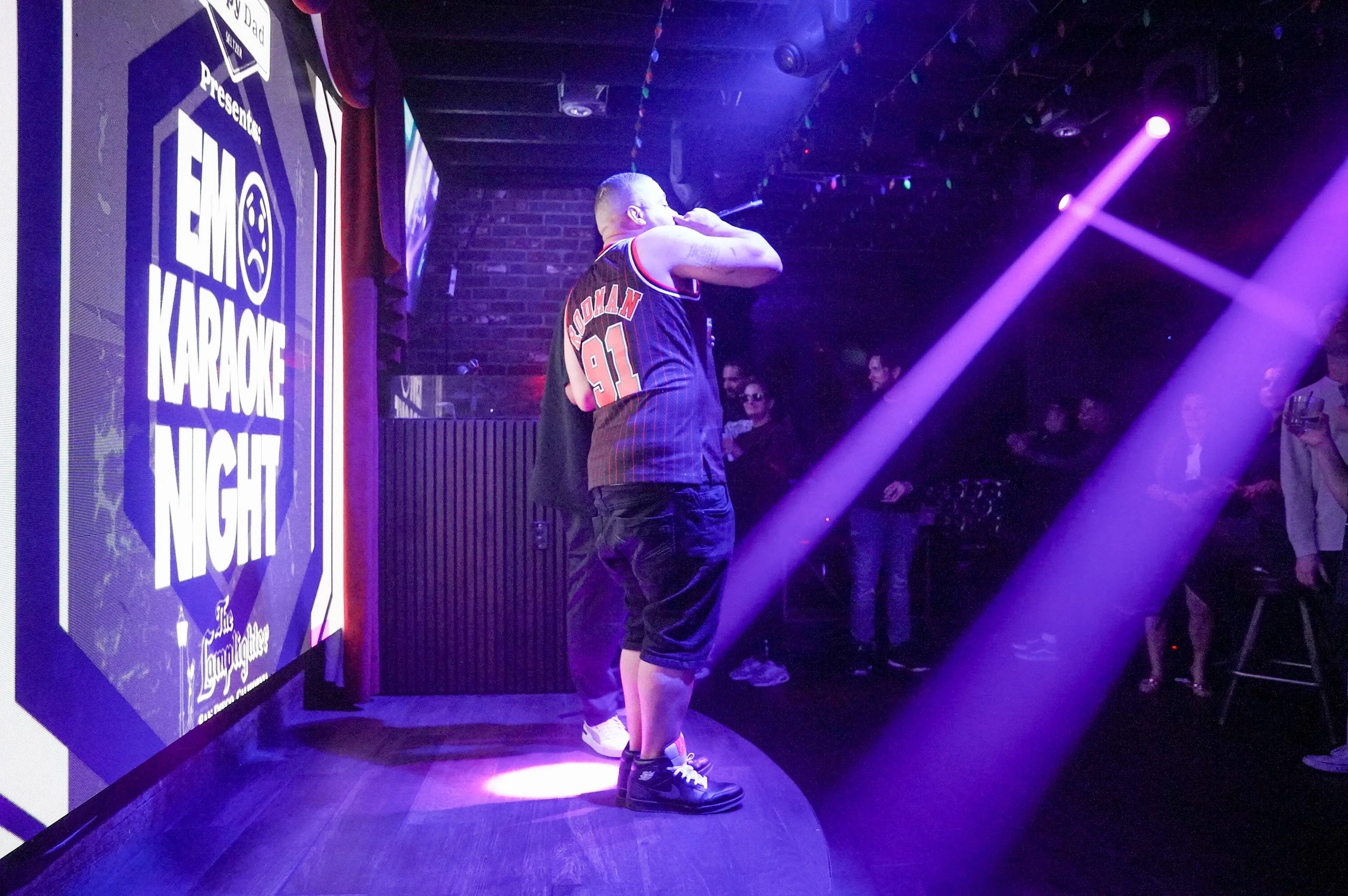 A man on stage during a karaoke night event, illuminated by purple stage lights, with a large sign that reads 'Emo Karaoke Night' on the left side, and a small crowd watching in the background.
