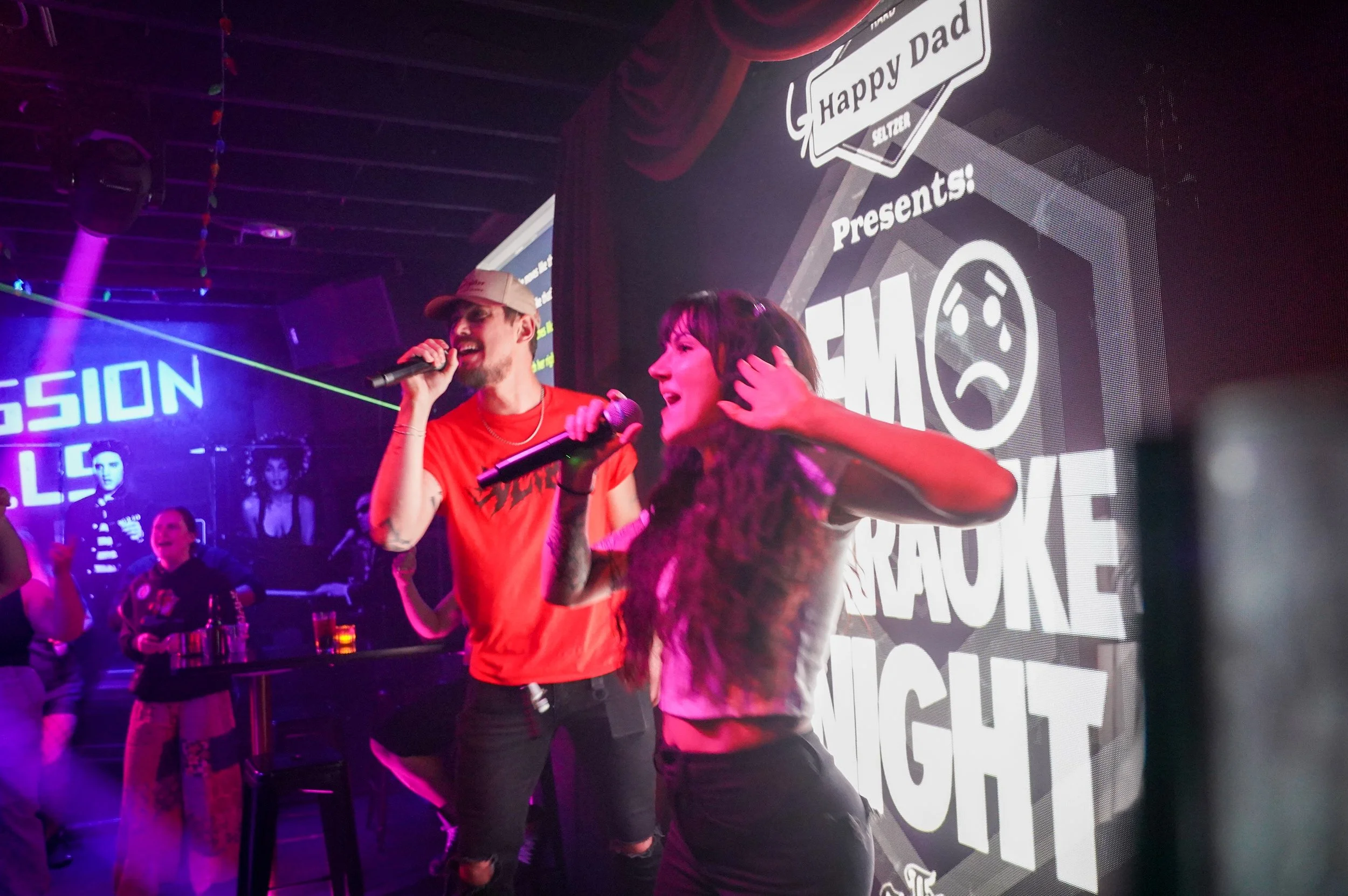 Two people singing karaoke on stage with microphones, a man in a red shirt and a woman with long hair, at a music night event with a large screen behind them showing promotional text and images.