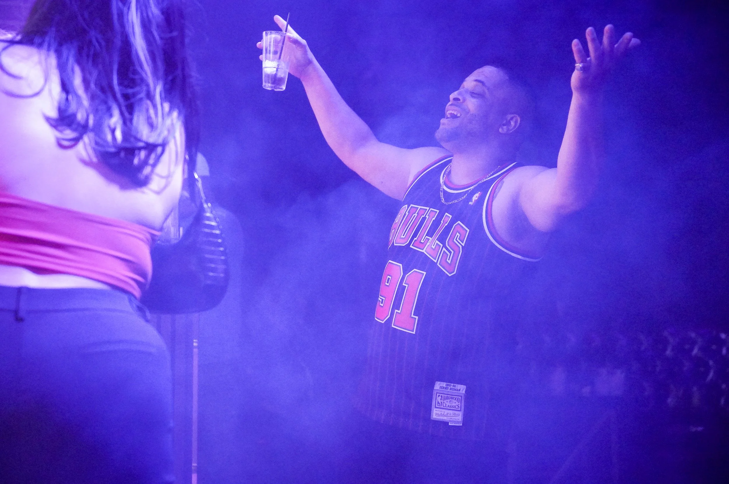 A man smiling and dancing with his arms raised, wearing a Chicago Bulls jersey, holding a drink, in a smoky purple-lit environment.