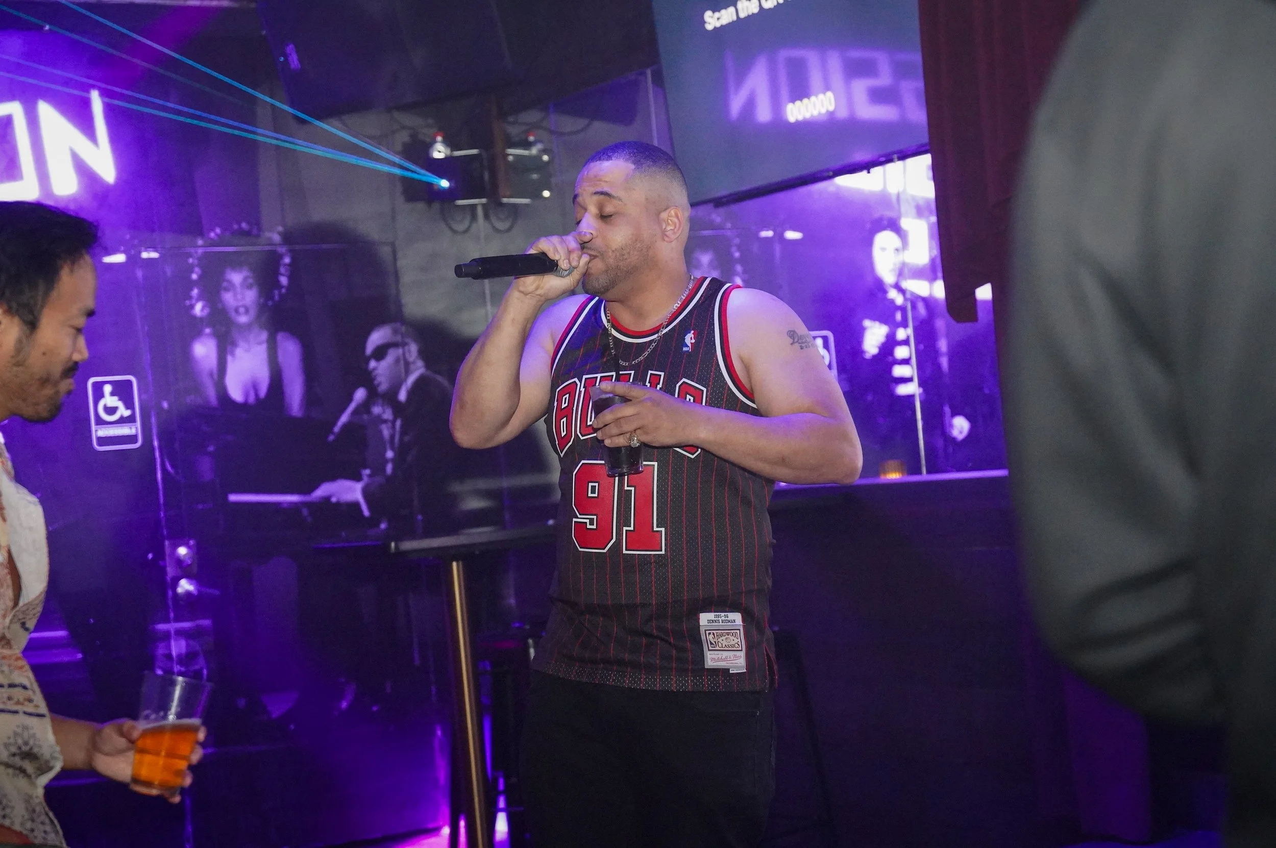A man wearing a Chicago Bulls basketball jersey singing into a microphone at a karaoke bar with purple lighting.