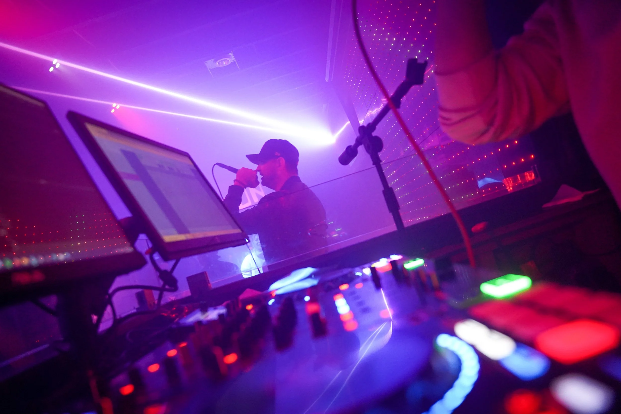 A DJ performs in a nightclub with colorful LED lights, a computer, and a microphone, while a person in the foreground watches.