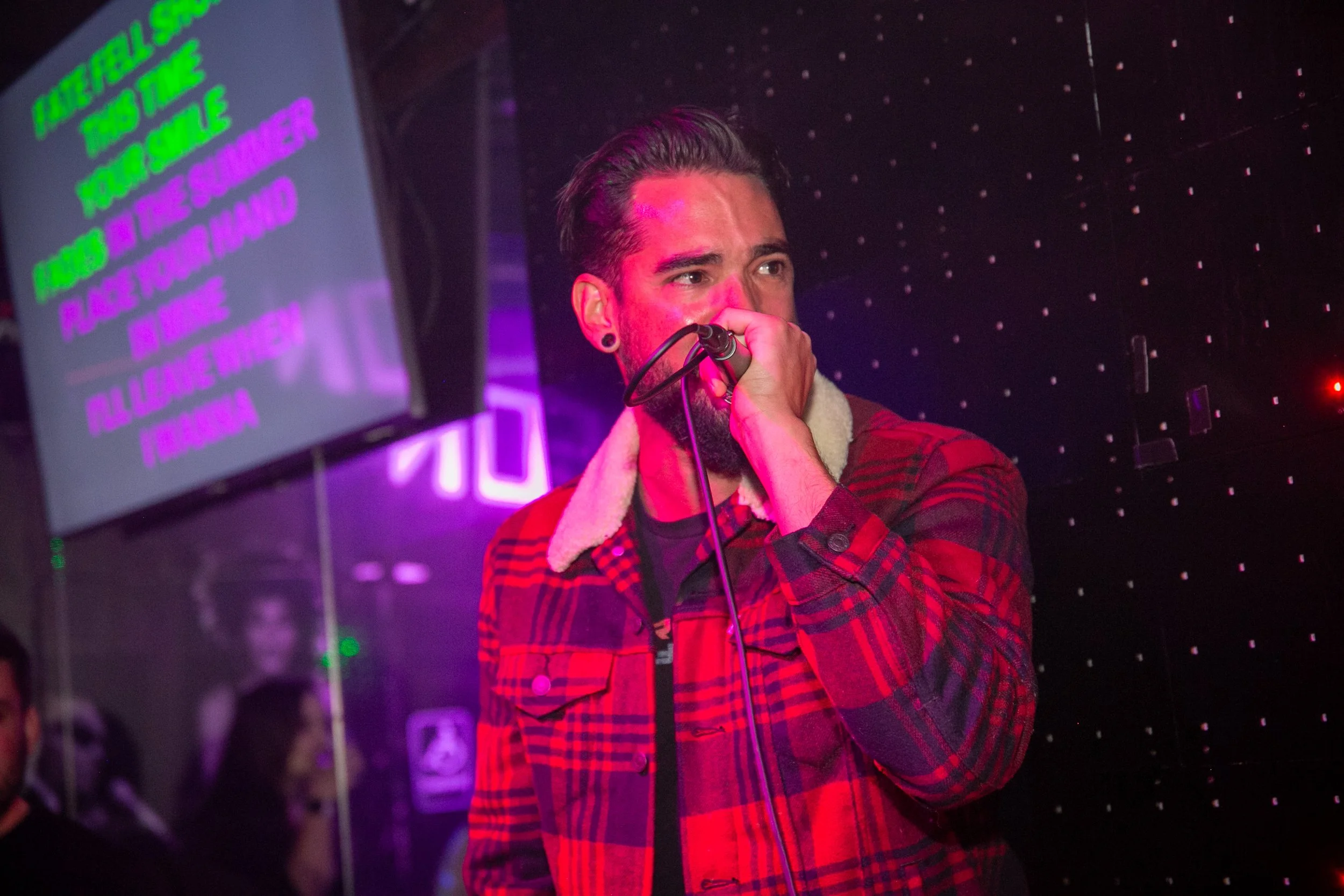 A man holding a microphone, singing or speaking, with a serious expression, wearing a red and black plaid shirt with a shearling collar, in a room with neon and purple lighting, and a screen displaying colorful text.