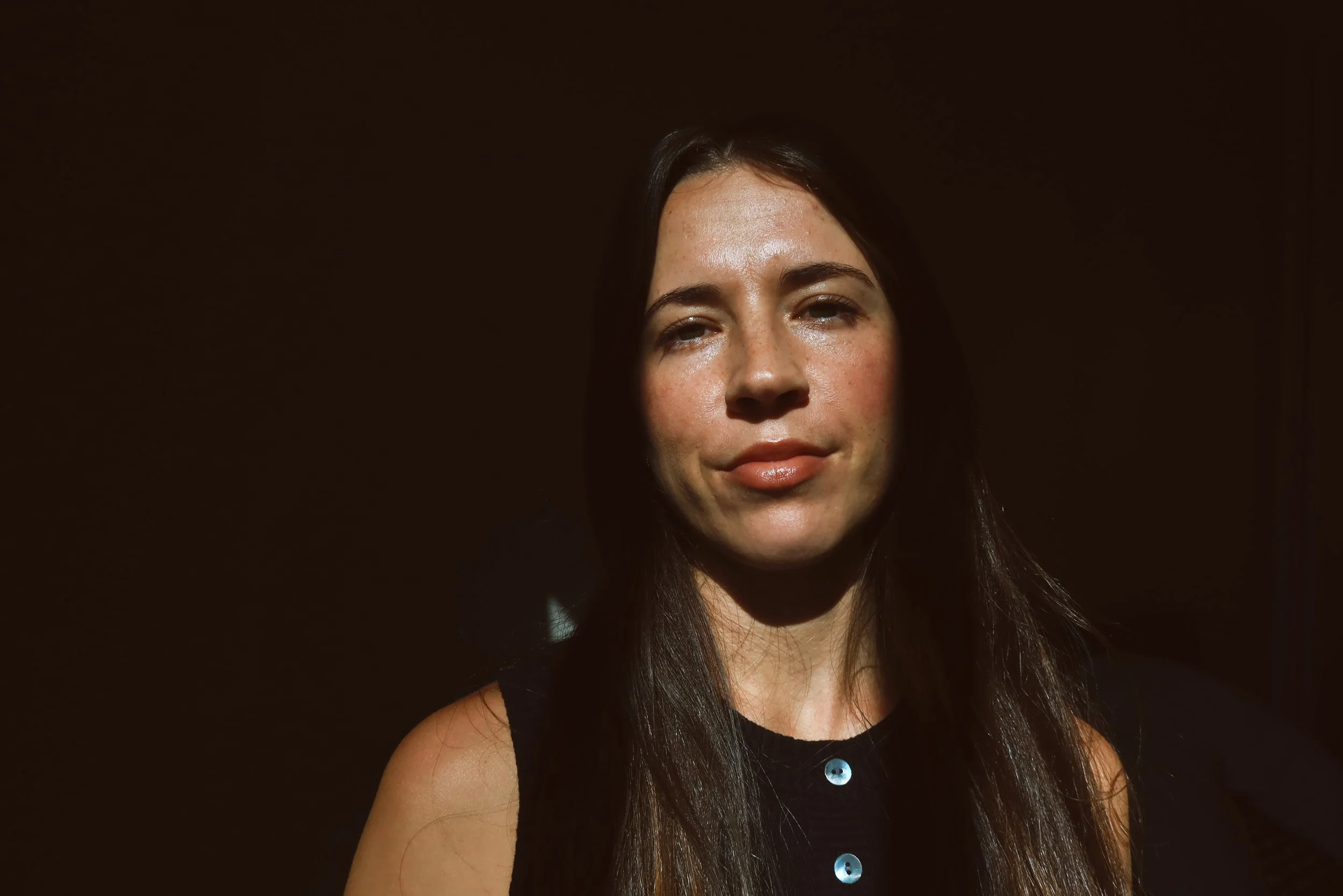 A woman with long dark hair takes in sunlight against a dark background, with some freckles on her face and wearing a sleeveless black top.