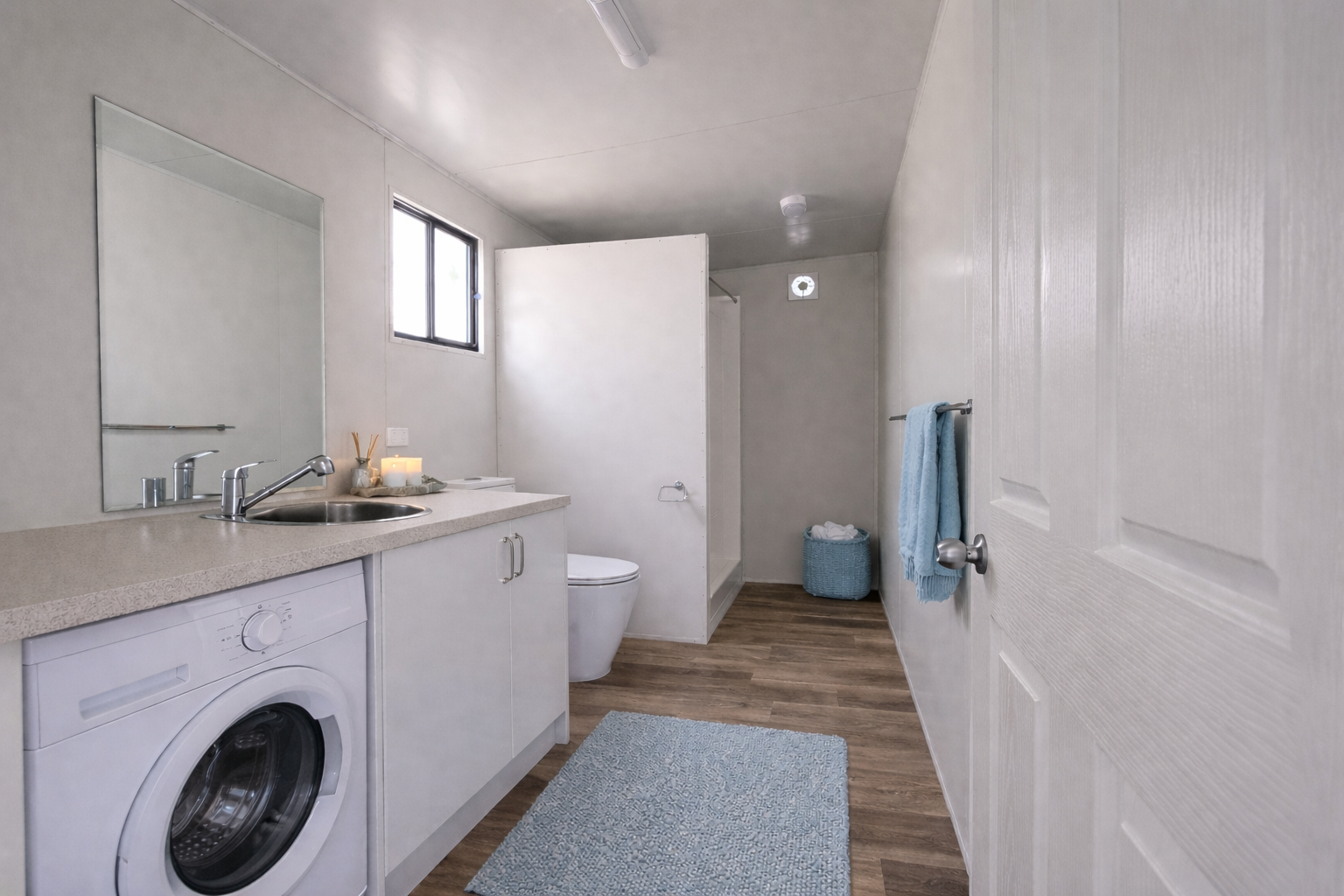 Interior of a bathroom with a washing machine, sink, mirror, toilet, window, towel rack with blue towel, laundry basket with white towels, and a light-colored rug on wood flooring.