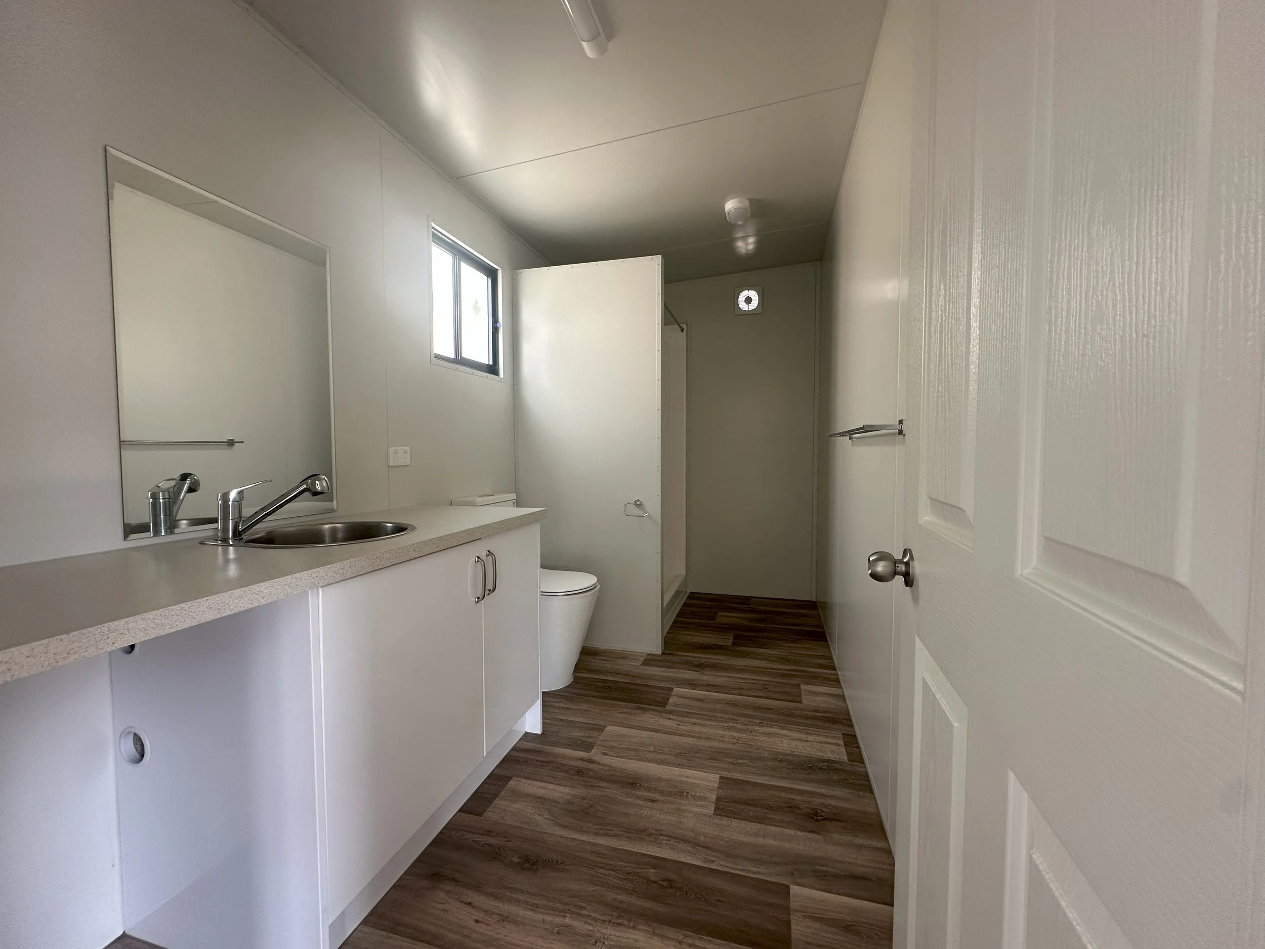 Empty bathroom with white cabinets, a sink, a mirror, a toilet, a small window, and light-colored wooden flooring.