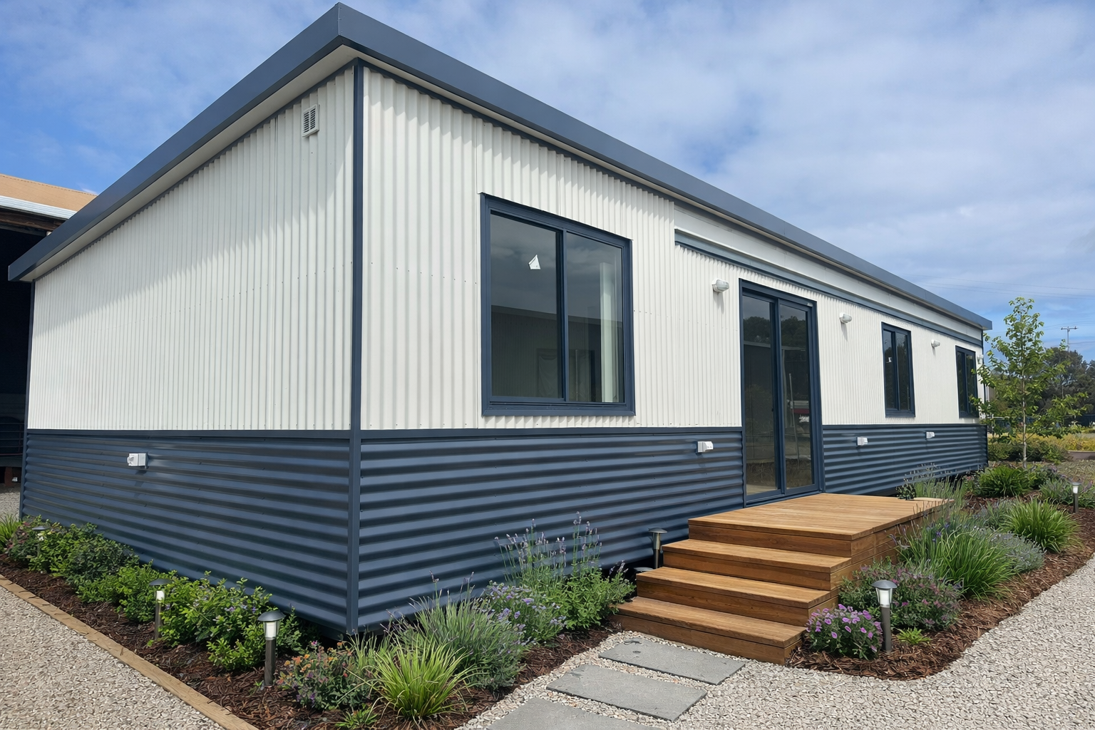 Modern tiny house with a white upper exterior and dark blue lower siding, large windows, wooden steps leading to a small porch, landscaped garden with greenery and flowers, gravel path, and a partly cloudy sky.