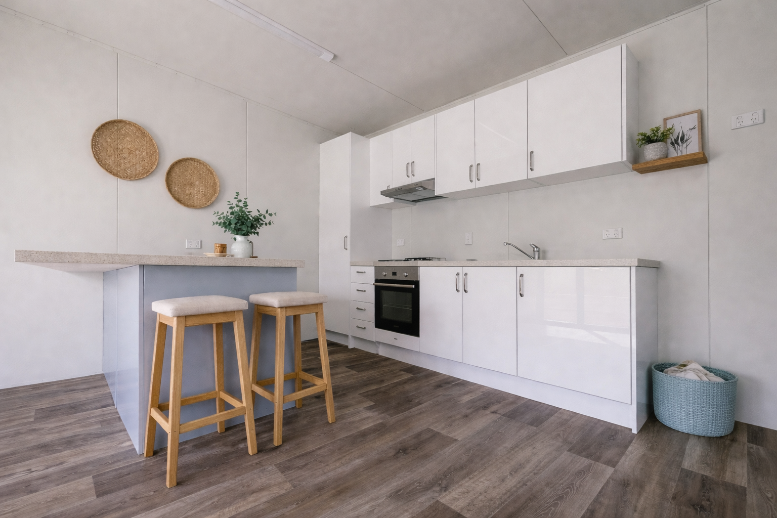Minimalist kitchen with white cabinets, a built-in oven, and a small counter with two wooden stools. Decor includes woven wall hangings, potted plants, and a teal storage basket on the floor.