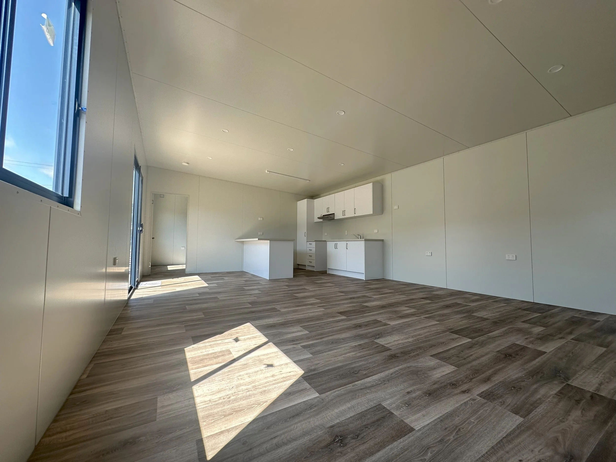An empty, modern, unfurnished kitchen with white walls and cabinets, wooden flooring, and a large window letting in natural light.