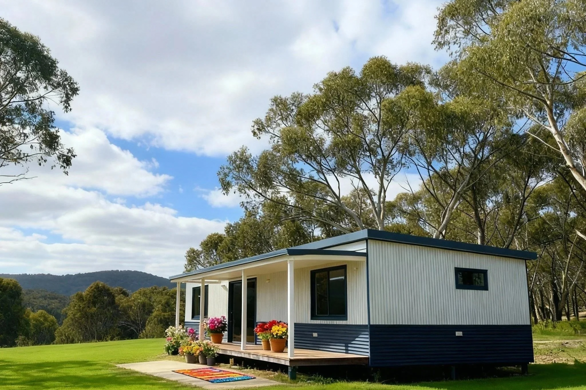 A small white house with a blue roof, wooden porch, and potted flowers in front, set in a green grassy area with trees and hills under a partly cloudy sky.