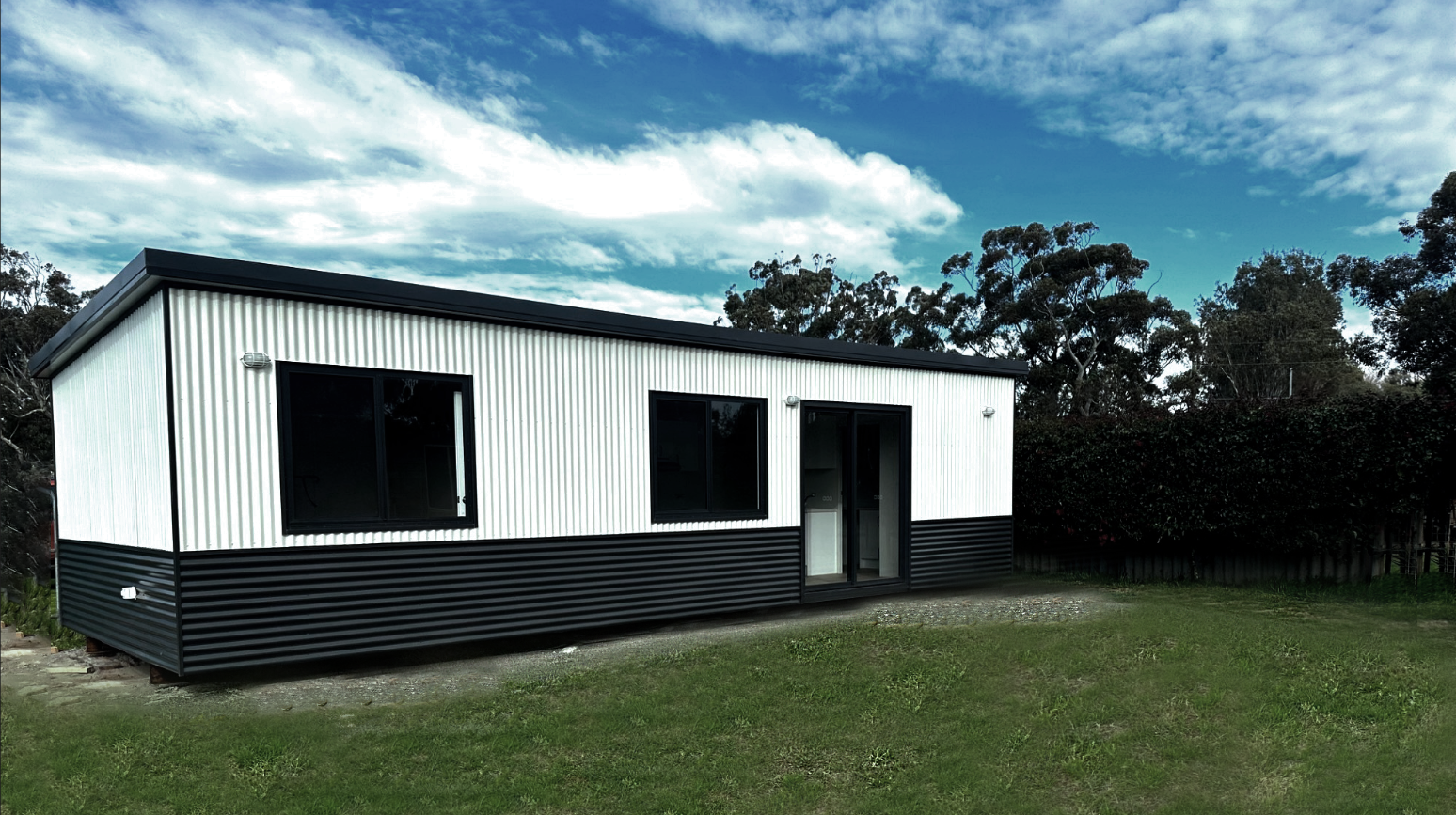 Modern tiny house with white and black exterior siding, large sliding glass doors and windows, situated on a grassy lawn with trees and cloudy sky in the background.