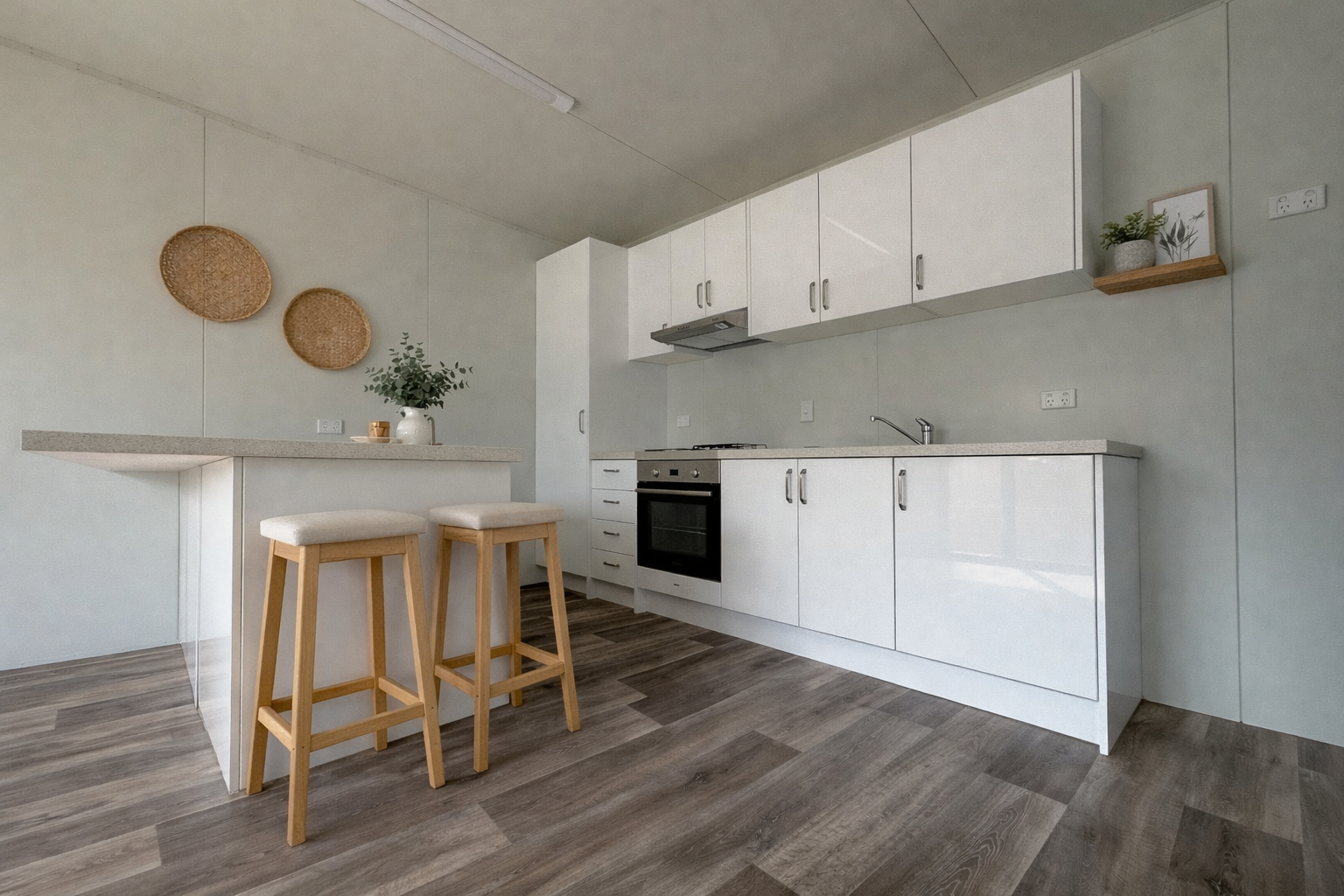 Minimalist kitchen with white cabinets and countertops, a built-in stainless steel oven, a sink, and a small breakfast bar with two wooden stools, decorated with wall baskets and potted plants.