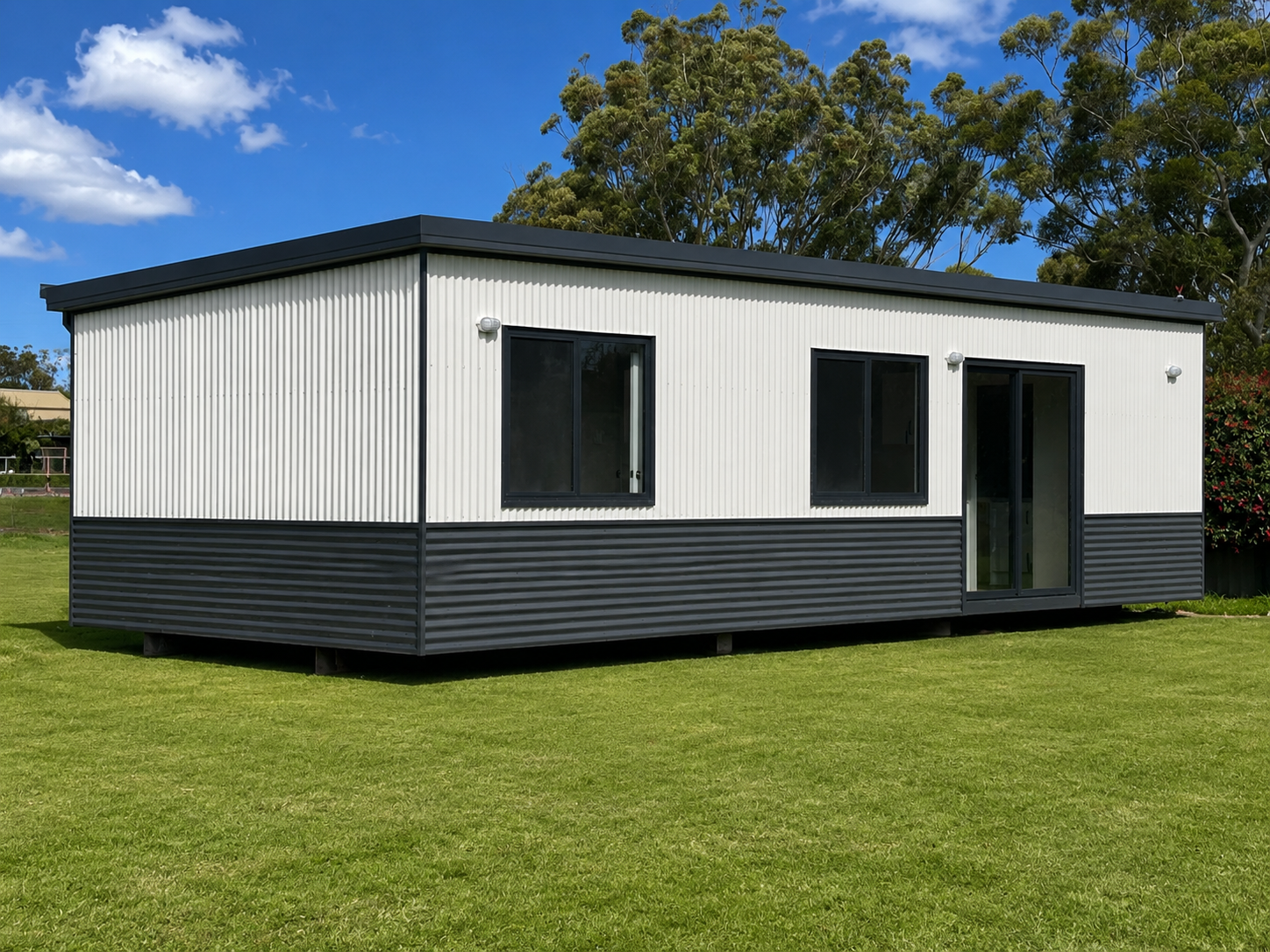 A modern tiny house with white vertical siding and black trim, situated on a well-maintained grassy yard under a blue sky with trees in the background.