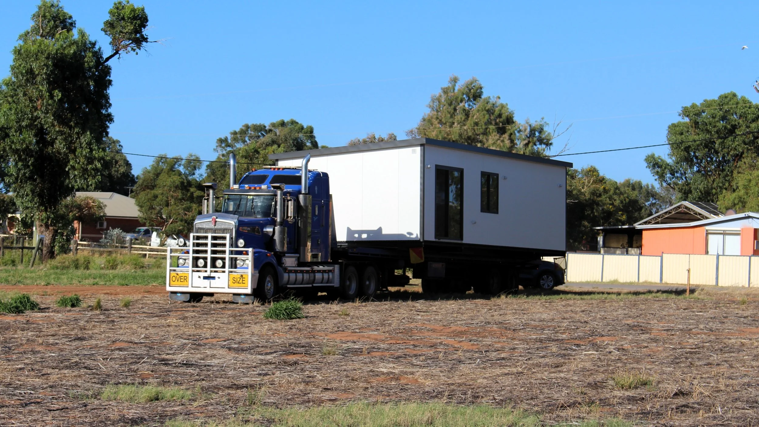 A blue semi-truck with a white container is parked on a partially cleared dirt lot, surrounded by trees and residential buildings, under a bright blue sky.