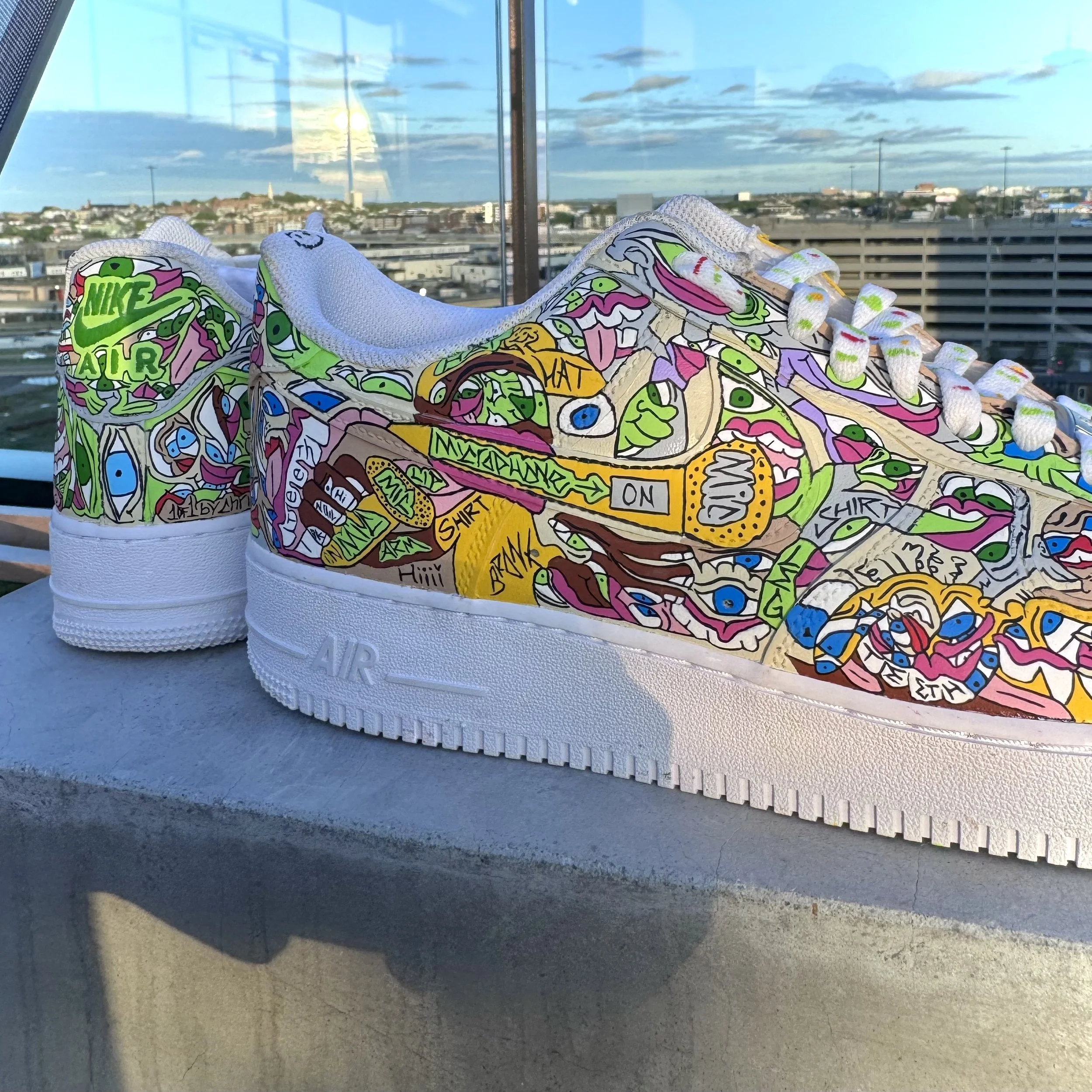 Colorful, graffiti-style design on a pair of white Nike Air Force sneakers, featuring cartoonish faces, eyes, and various words and phrases, with a cityscape and sky in the background.