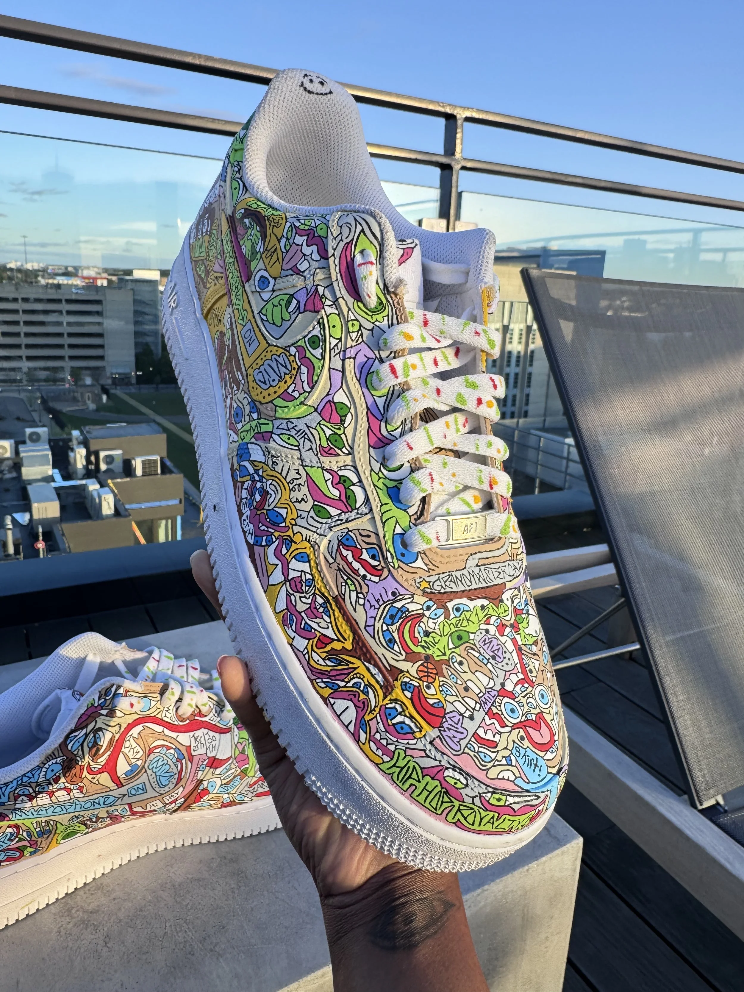 Close-up of a colorful, graffiti-style decorated sneaker being held outdoors on a rooftop, with a cityscape and clear sky in the background.