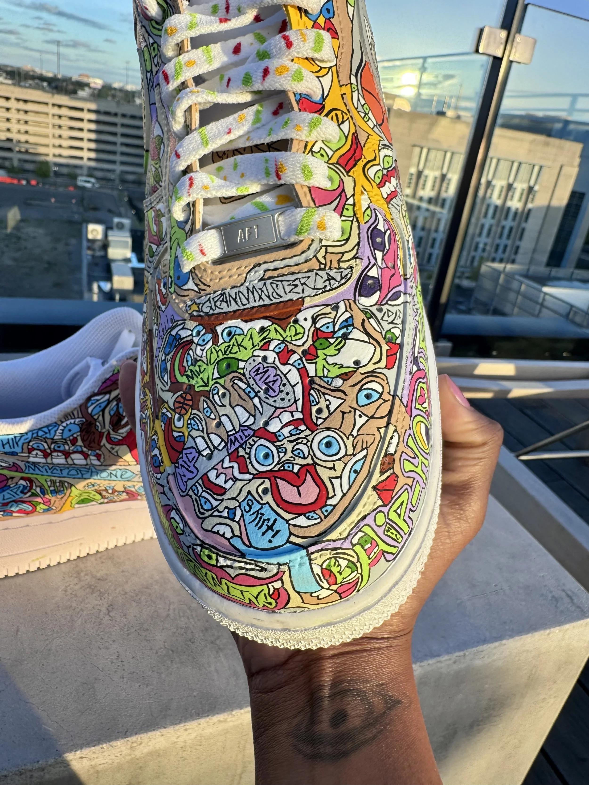 Close-up of a colorful, hand-drawn decorated sneaker with various doodles, characters, and words, held against a cityscape background during sunset.