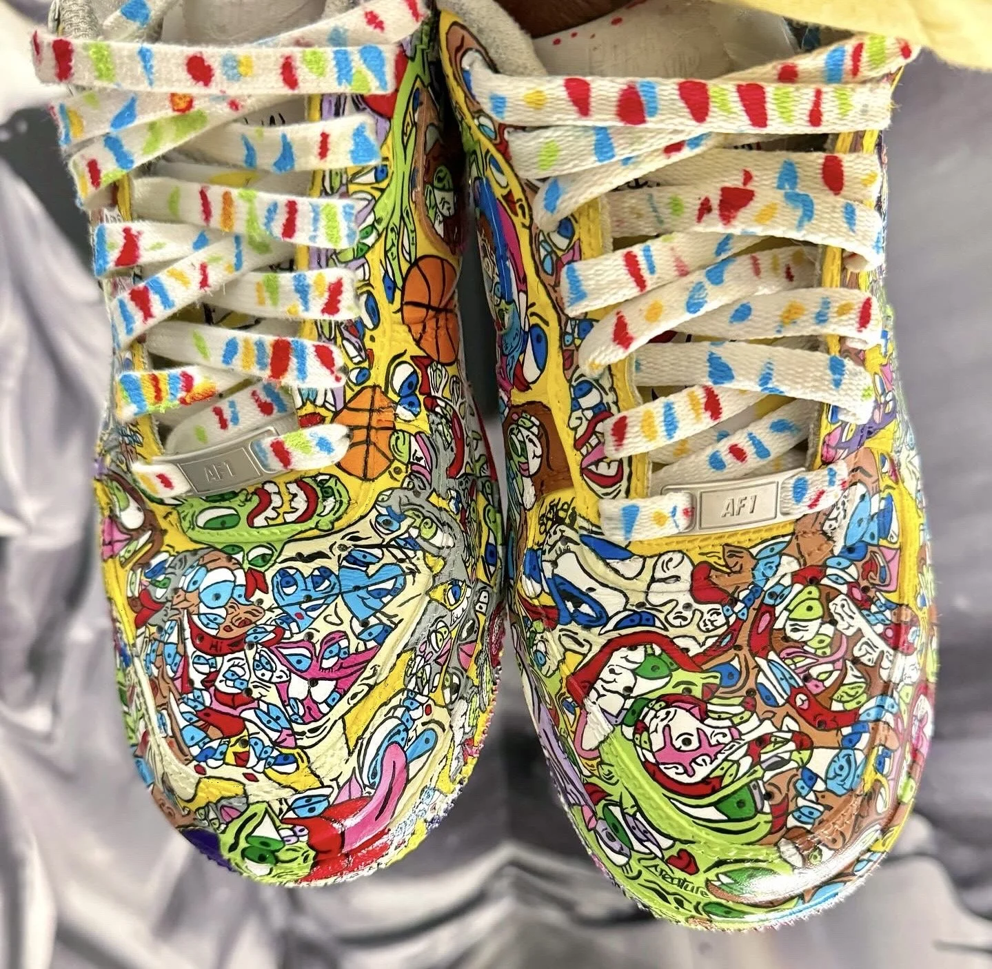 Colorful sneakers with graffiti-style artwork featuring various abstract characters and designs, laces with rainbow-like patterns.