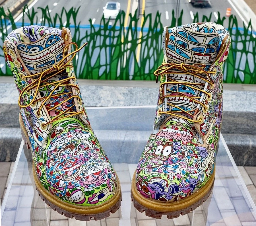 Colorful, graffiti-style decorated boots with various cartoonish and comic characters, placed on a clear display stand.