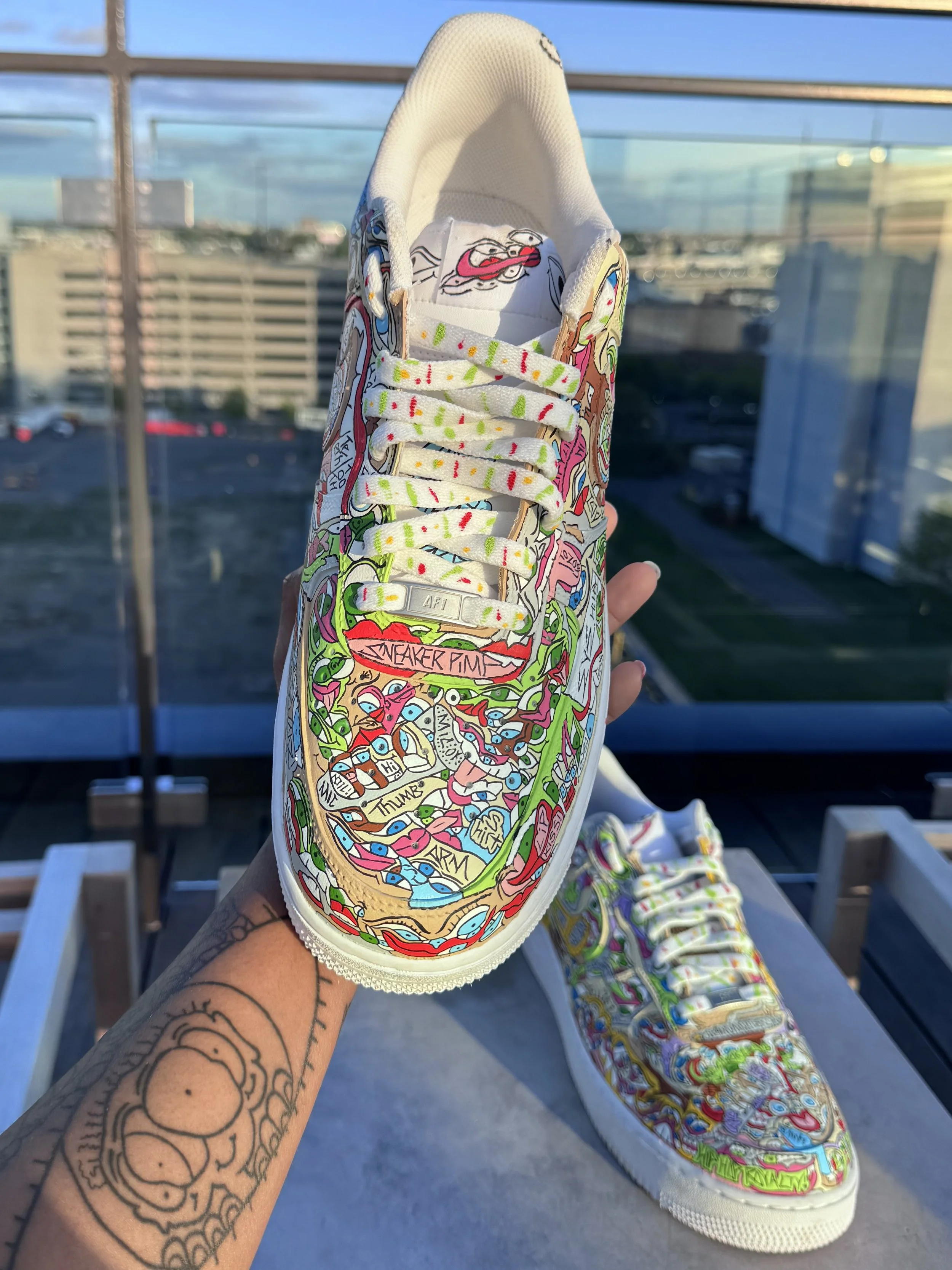 A colorful sneaker with graffiti-style artwork covering its surface, held in a person's hand with a tattoo on their wrist.