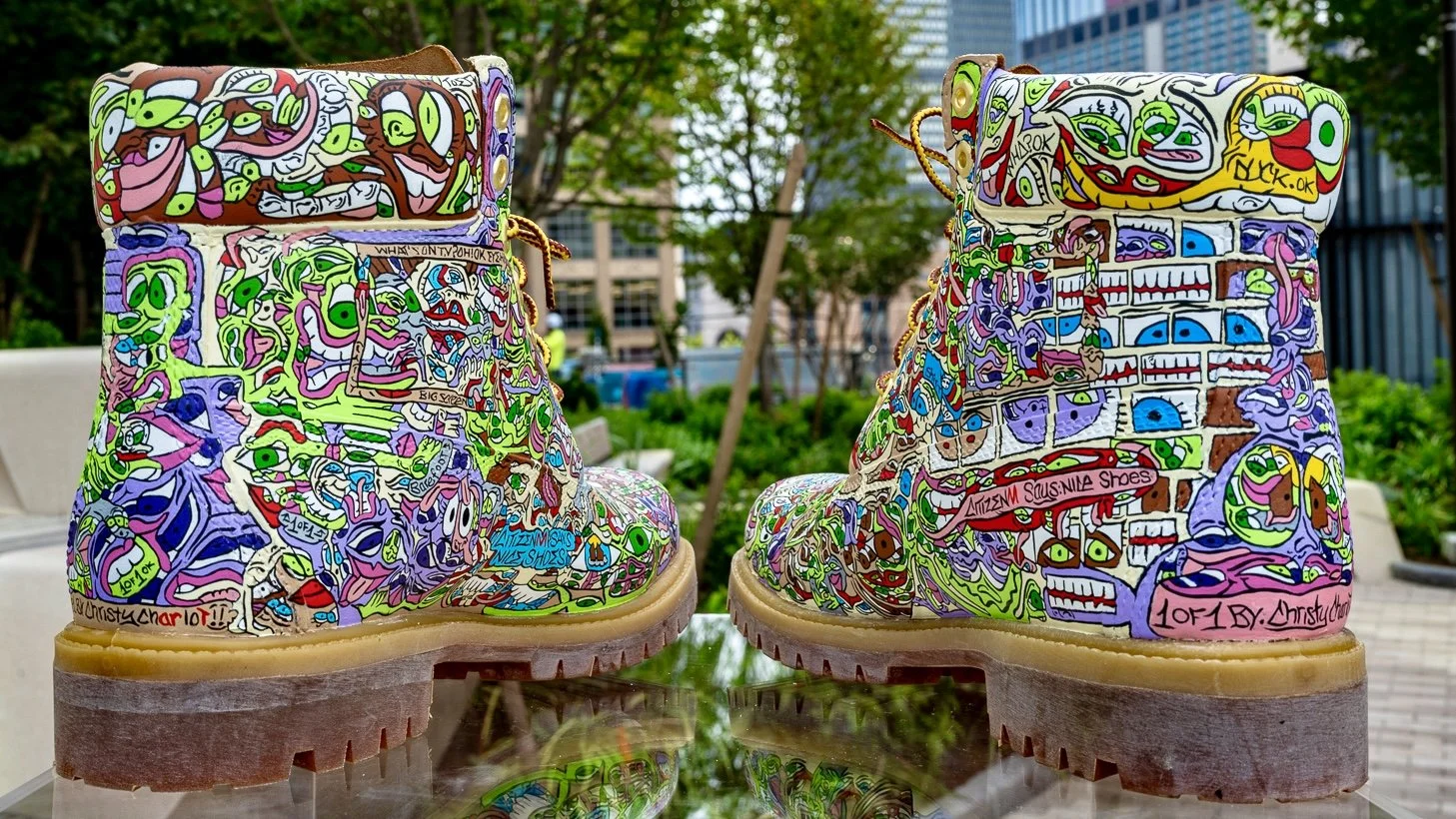 A pair of boots painted with colorful, abstract, doodle-style artwork featuring various faces, eyes, and shapes, designed by Christy Charlotte.