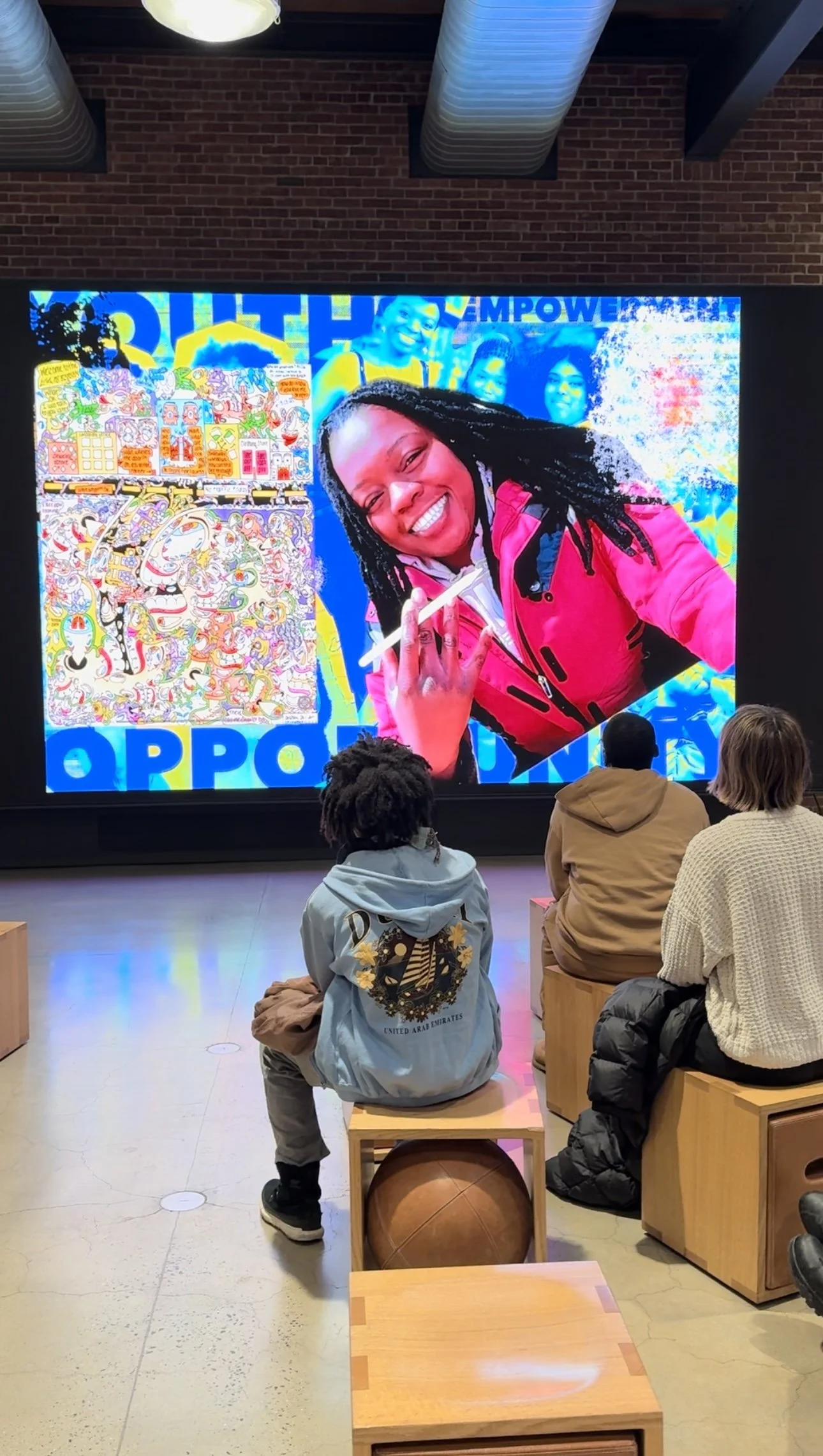 People sitting on wooden stools watching a large colorful digital screen displaying a woman smiling and holding a pen, with a background of vibrant artwork and faces.