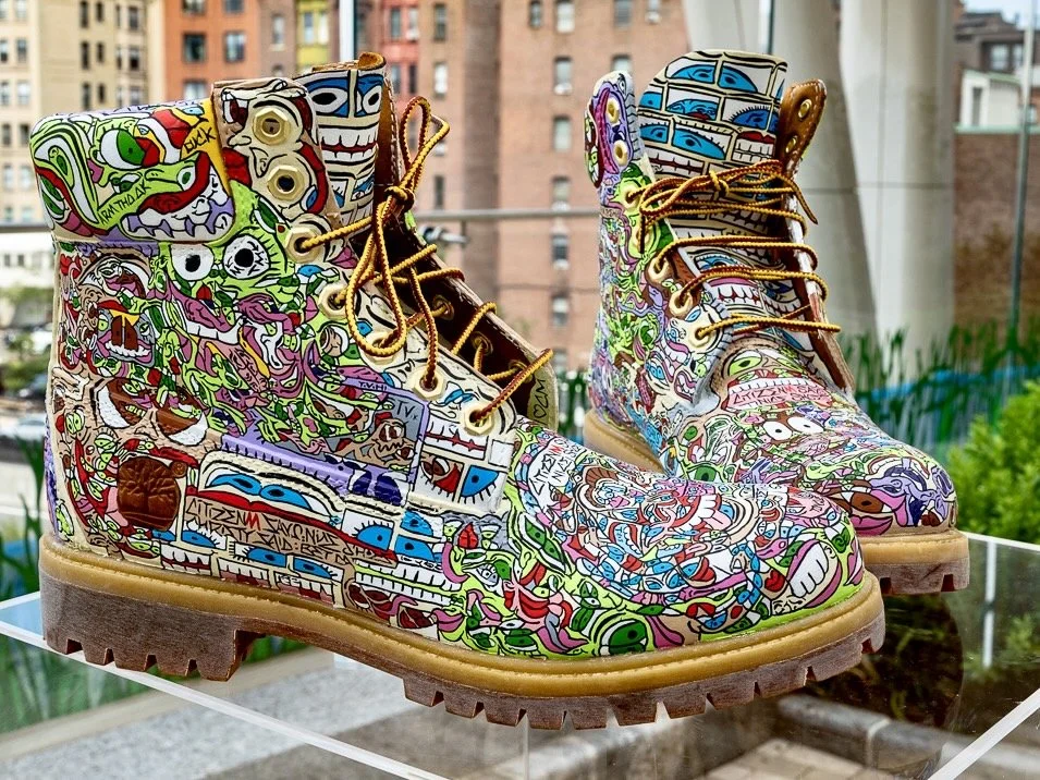 Colorful, illustrated high-top boots with various cartoon characters and graffiti art, displayed outdoors with city buildings in the background.
