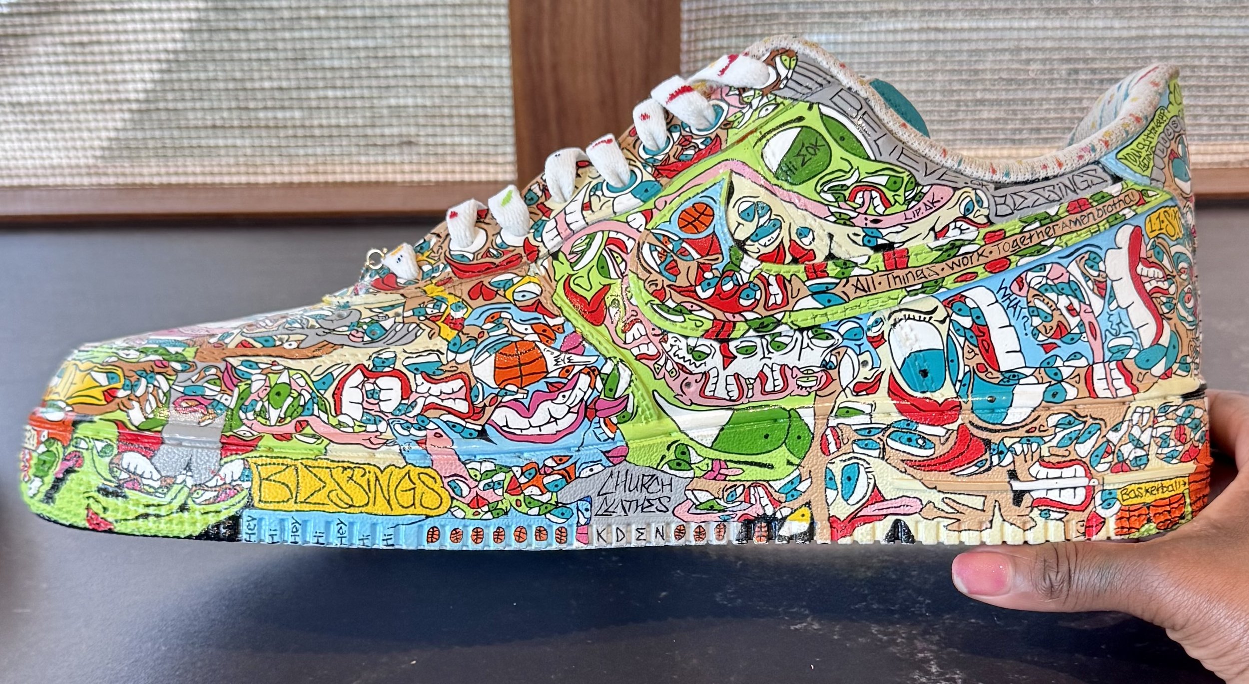 Colorful, graffiti-style sneaker with various cartoon characters, words, and patterns, being held by a person's hand.