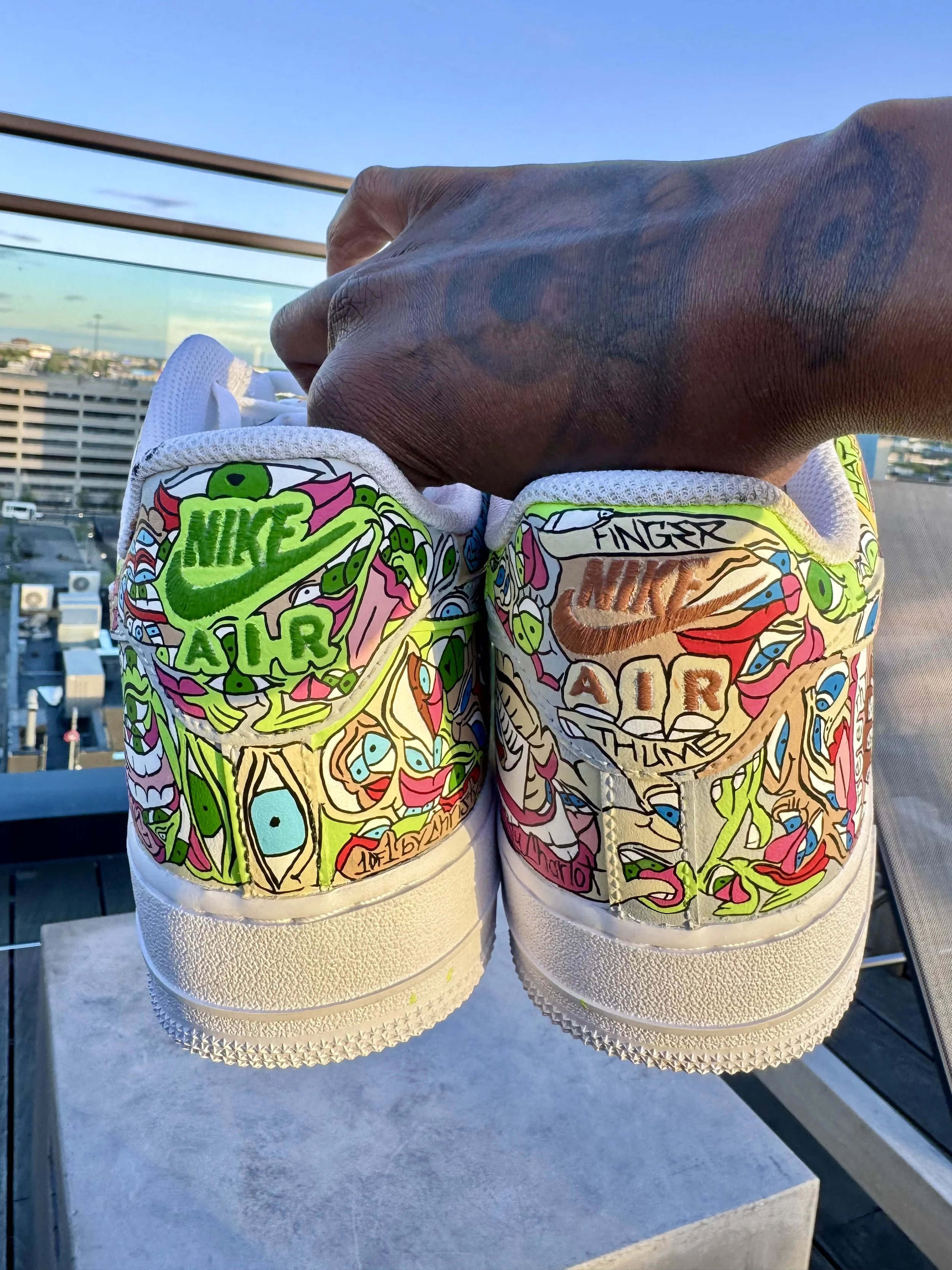 Close-up of hand holding two white Nike Air Force 1 sneakers with colorful, doodle-style artwork and inscriptions, against an outdoor cityscape background with buildings and a clear sky.