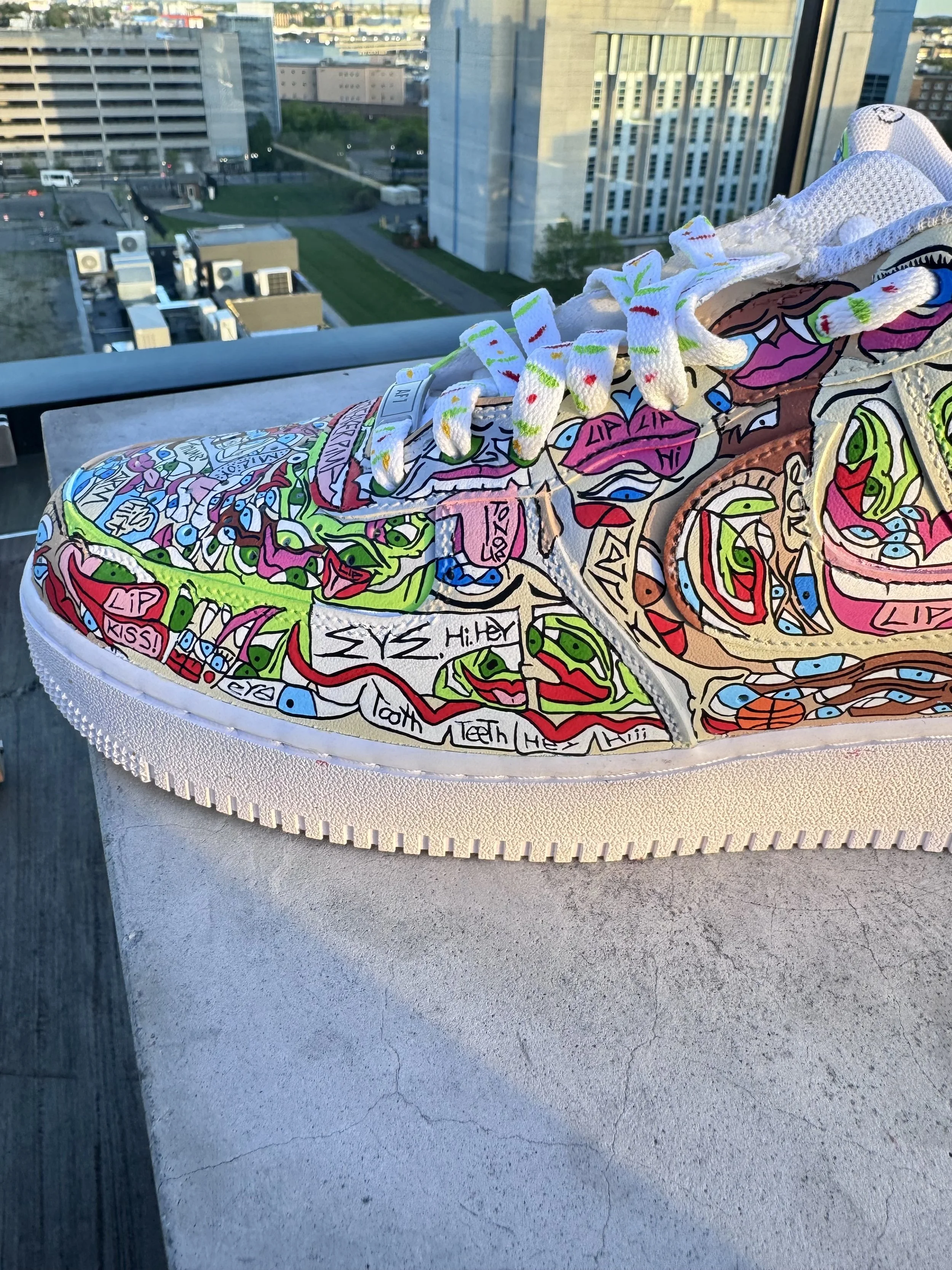 Colorful sneaker with graffiti-style art and handwritten messages, placed on a white surface with a city skyline in the background.