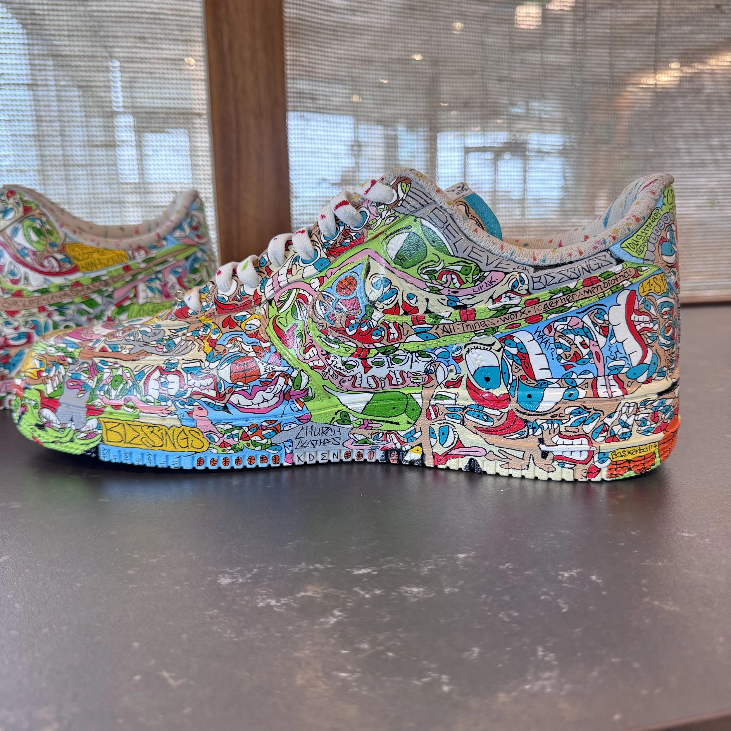Colorfully decorated Adidas sneaker with intricate, doodle-style artwork covering the entire shoe, placed on a dark surface with a blurred background of windows and wood paneling.
