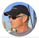 A person wearing sunglasses, a black hat, and a gray shirt, outdoors with blue skies.