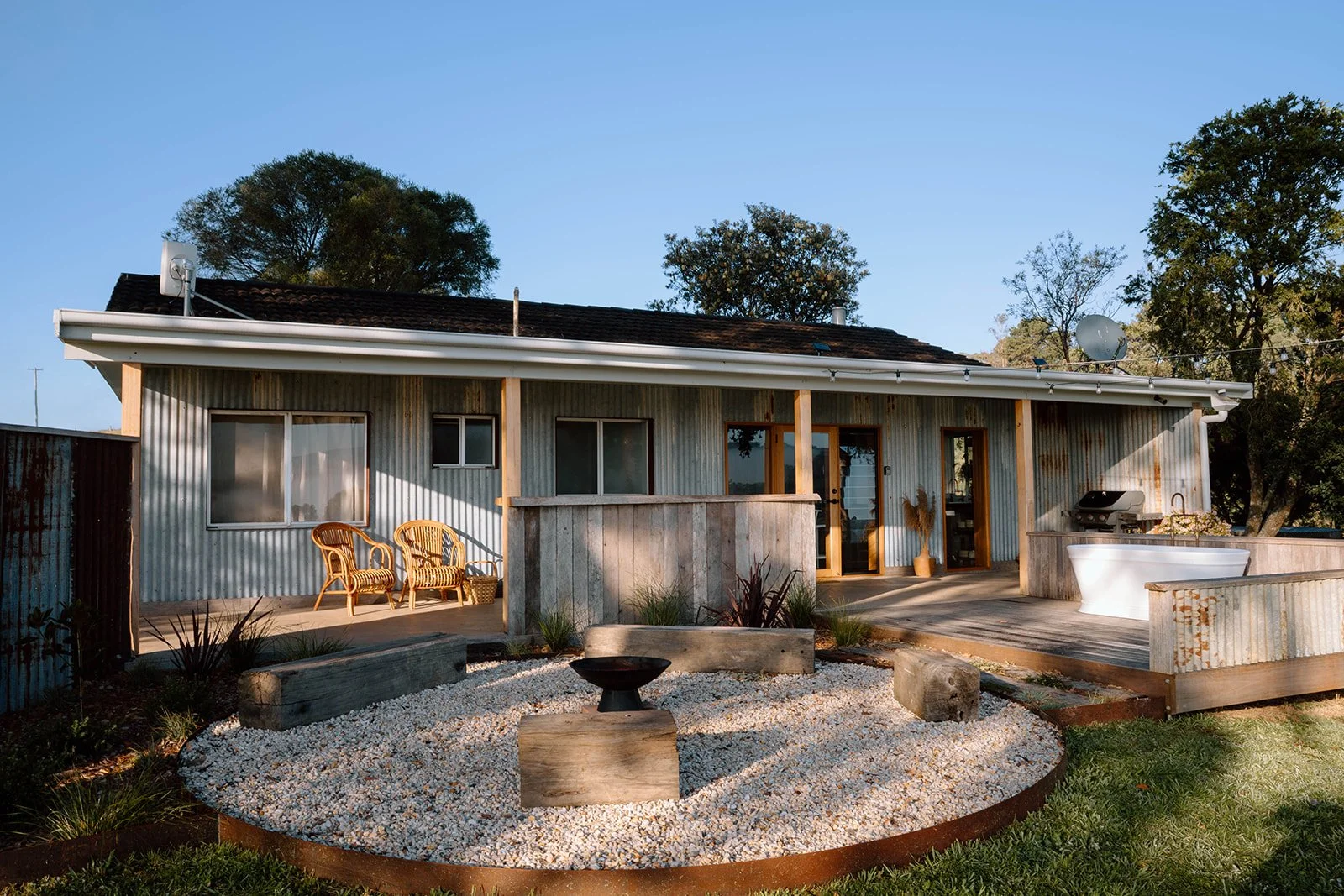 Backyard of a house with a corrugated metal exterior, wooden accents, and a deck with outdoor furniture, a grill, a bathtub, and decorative plants. There is a gravel garden area with rocks and outdoor art in the foreground, and trees in the backgroun