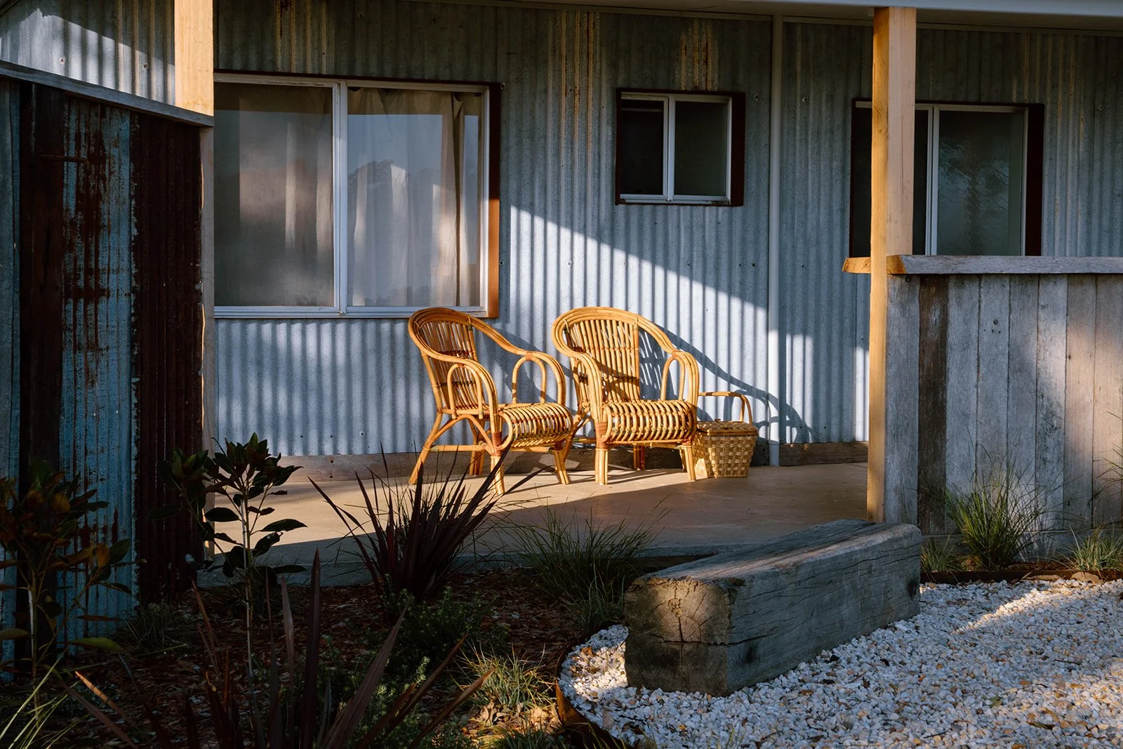 Two wicker chairs on a porch with a corrugated metal wall, plants, and gravel landscaping, illuminated by warm sunlight.