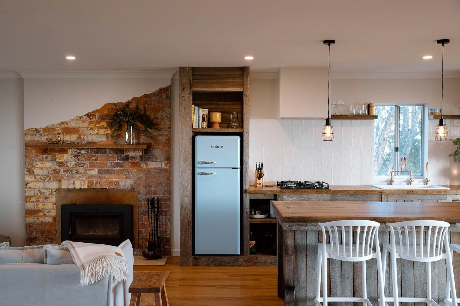 Open kitchen with exposed brick wall, rustic wood accents, a vintage blue refrigerator, white chairs, pendant lighting, and a window with trees outside.