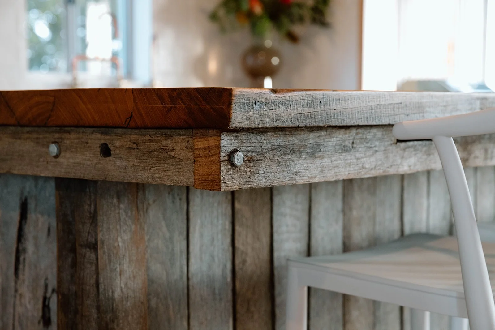 Close-up of a rustic wooden table corner with a weathered wood panel wall and a white chair in the background, with blurred window light.