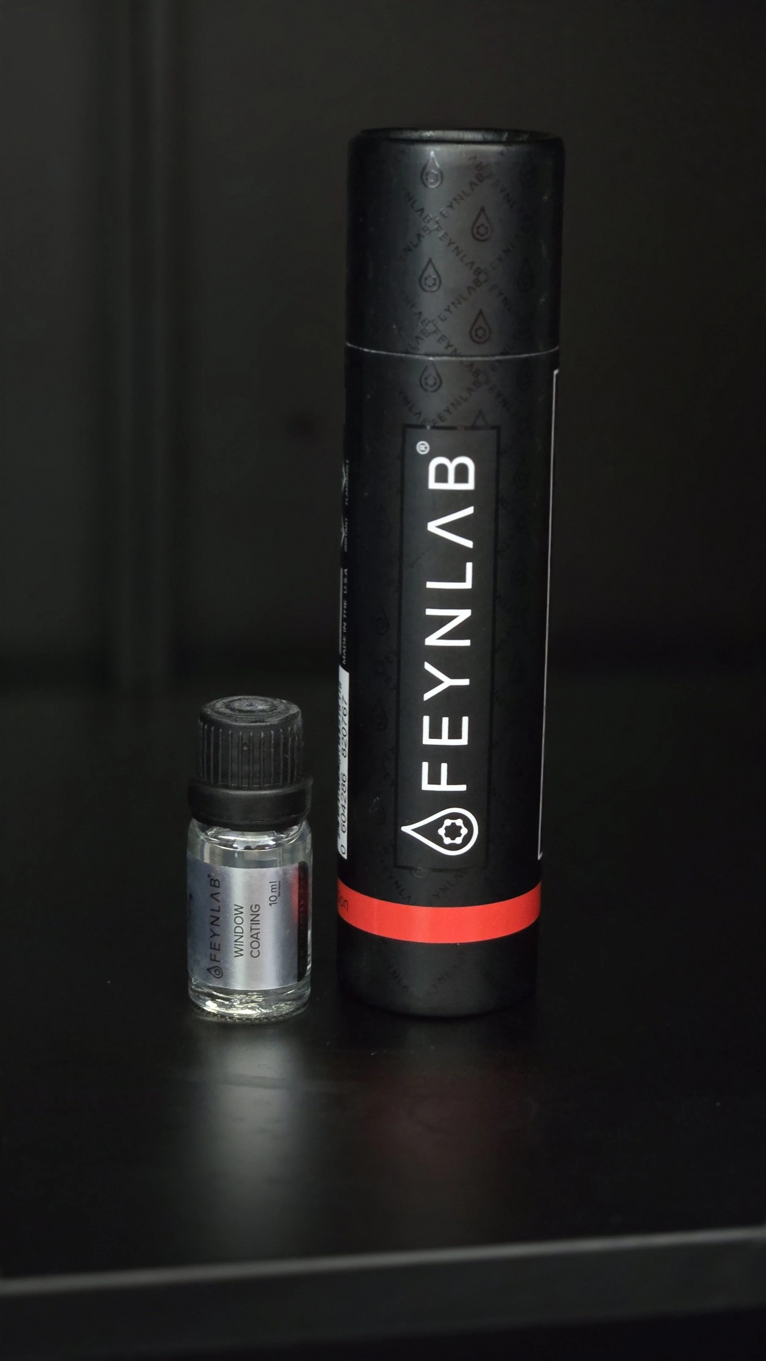 A small bottle labeled 'Feynlab Window Coating' and a larger black cylindrical container labeled 'Feynlab' sit on a black surface against a dark background.