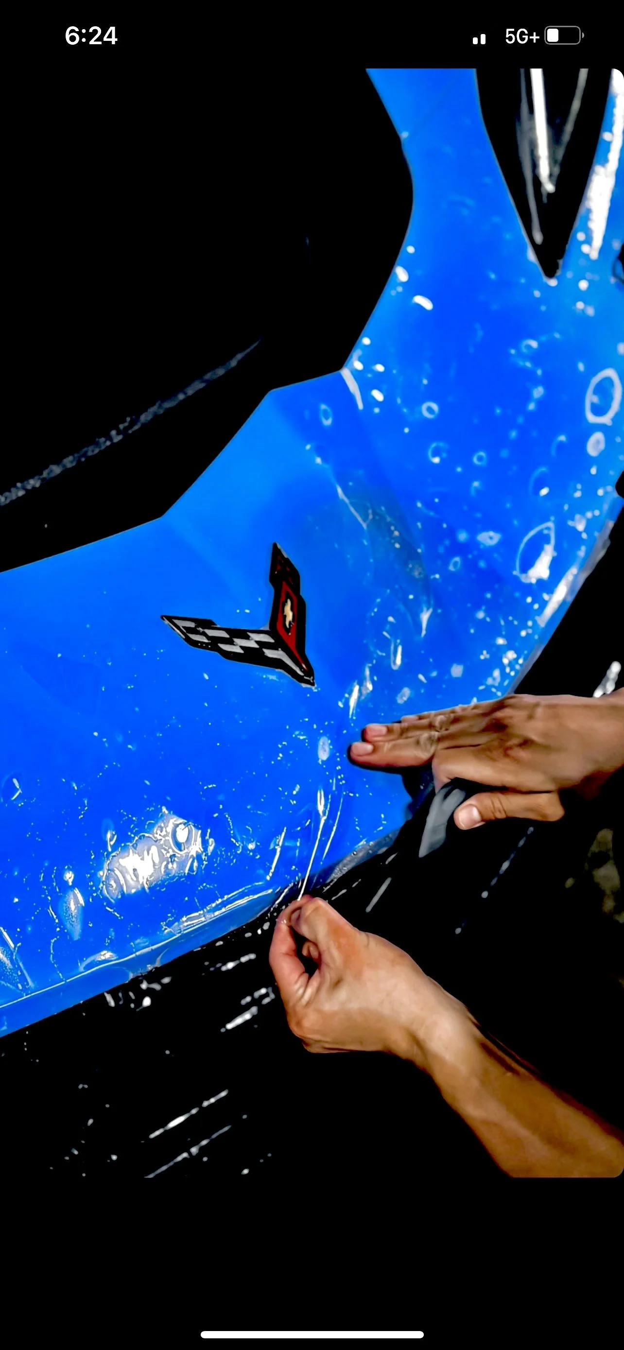 Person applying a vinyl decal with a Chevrolet Corvette logo onto a blue surface, surrounded by bubbles and water.