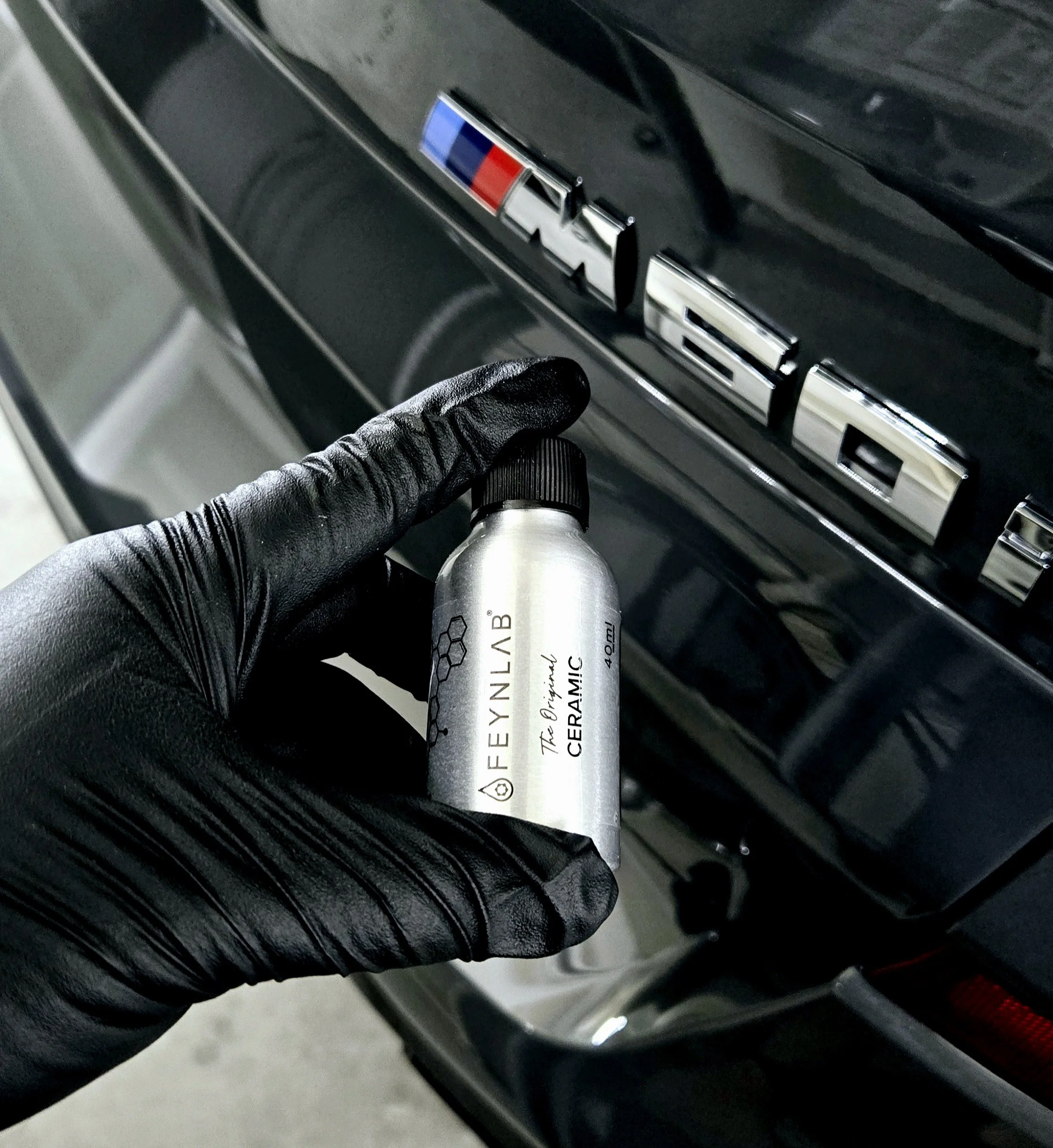 A person wearing a black glove holding a small bottle of Feynlab Ceramic spray in front of a black BMW with a M logo badge.