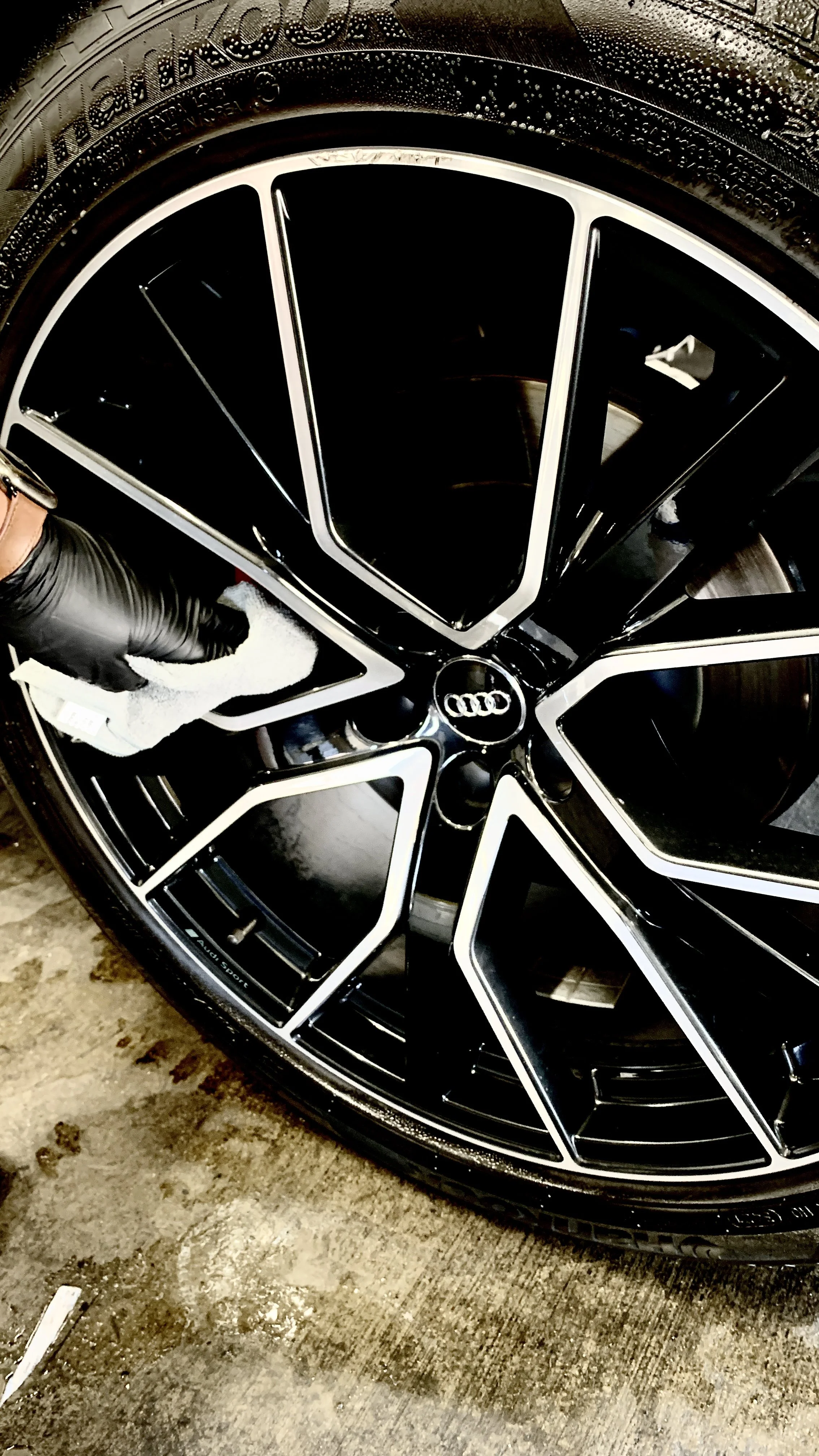 A person wearing a black glove polishing the black and silver alloy wheel of an Audi vehicle, with water droplets on the tire, on a concrete surface.
