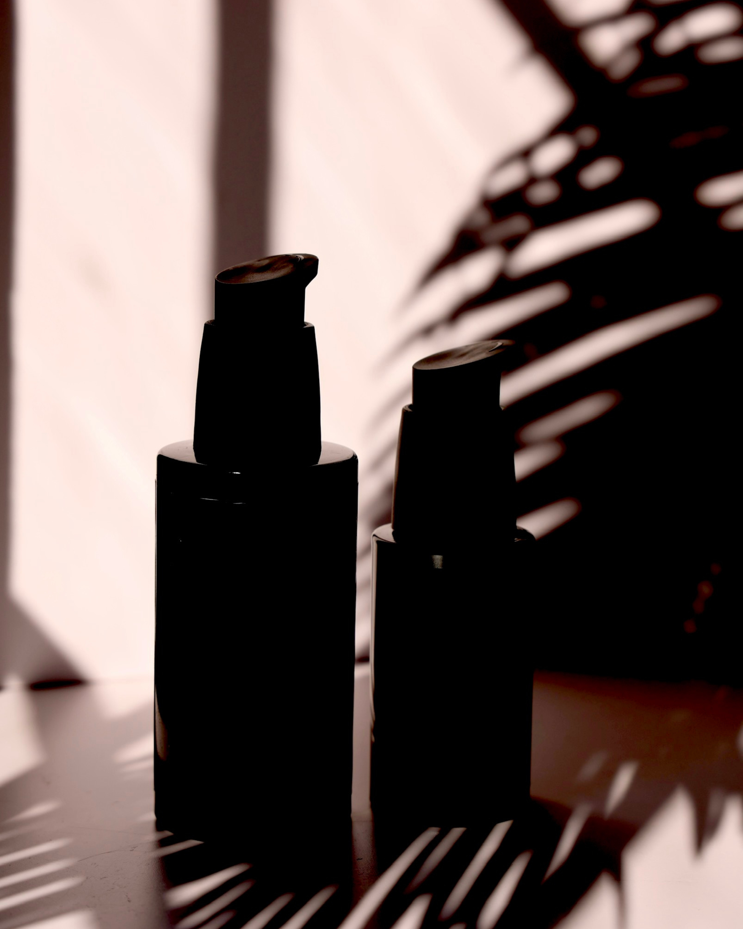 Silhouettes of two bottles with shadows and patterns from a nearby plant on a light background.