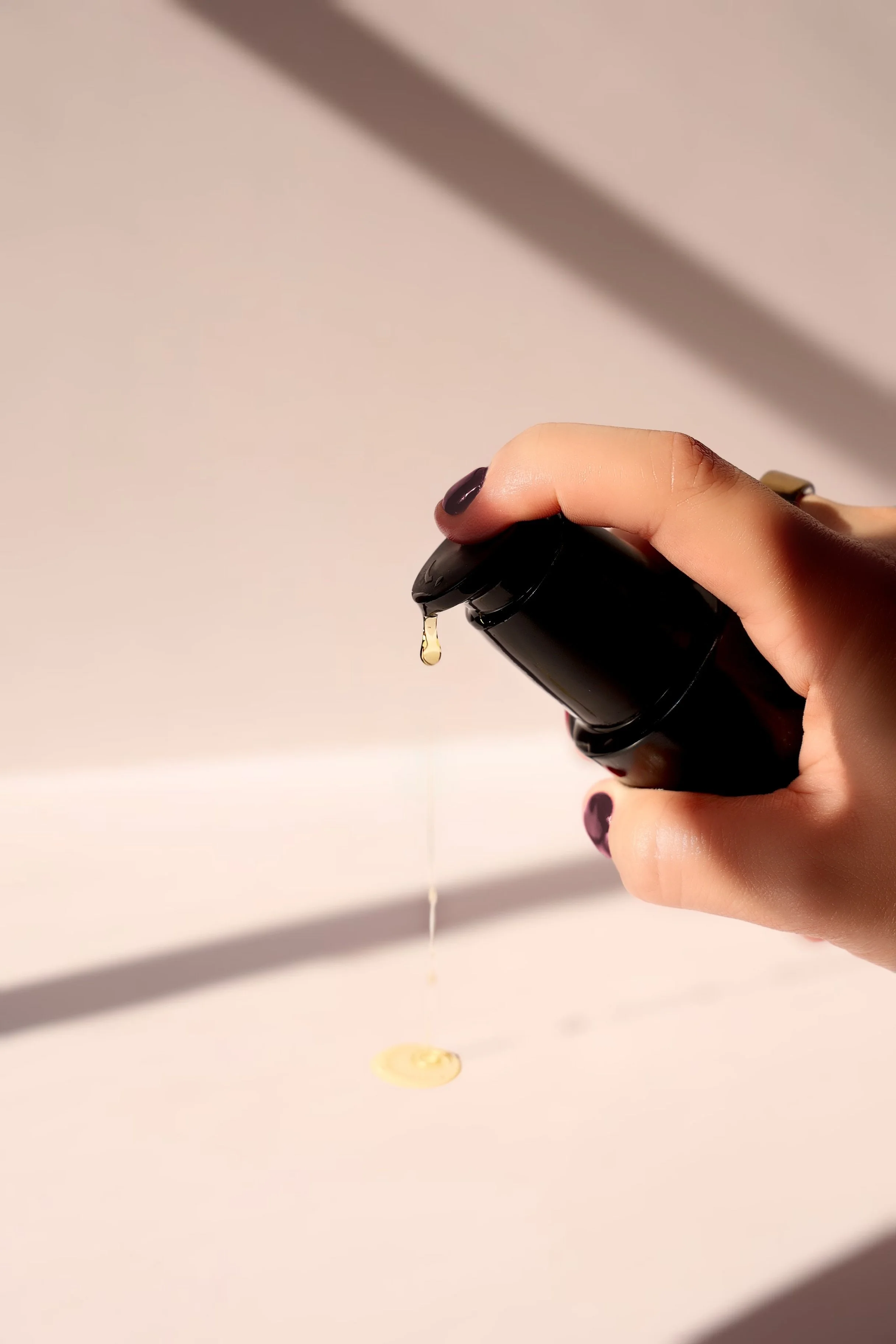 A hand with dark nail polish holding a black container, dispensing a drop of yellowish liquid onto a light-colored surface.