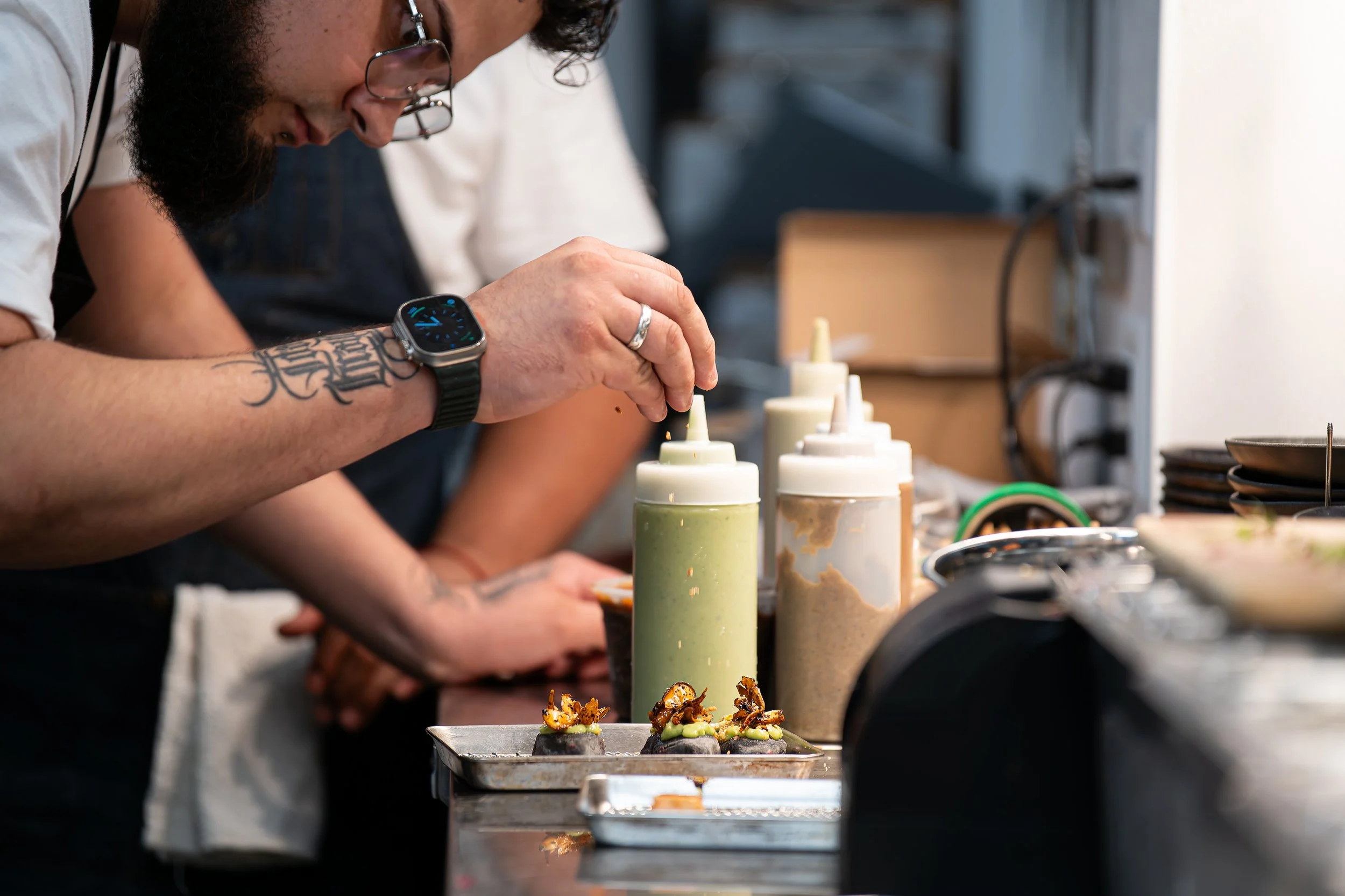 A chef decorates small plated dishes with sauce from squeeze bottles in a commercial kitchen.