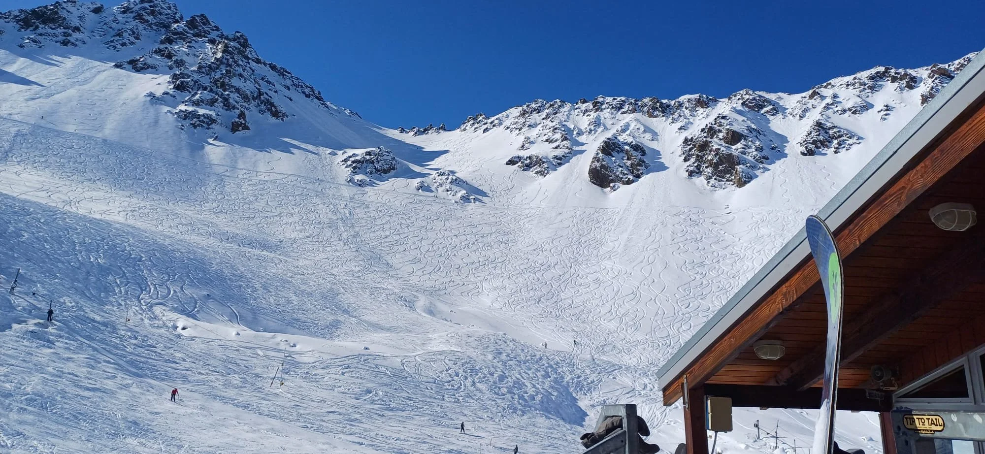 Snow-covered mountain slope with ski tracks, skiers, and a wooden building with a partial sign reading 'Tip to Taal' on the right side.